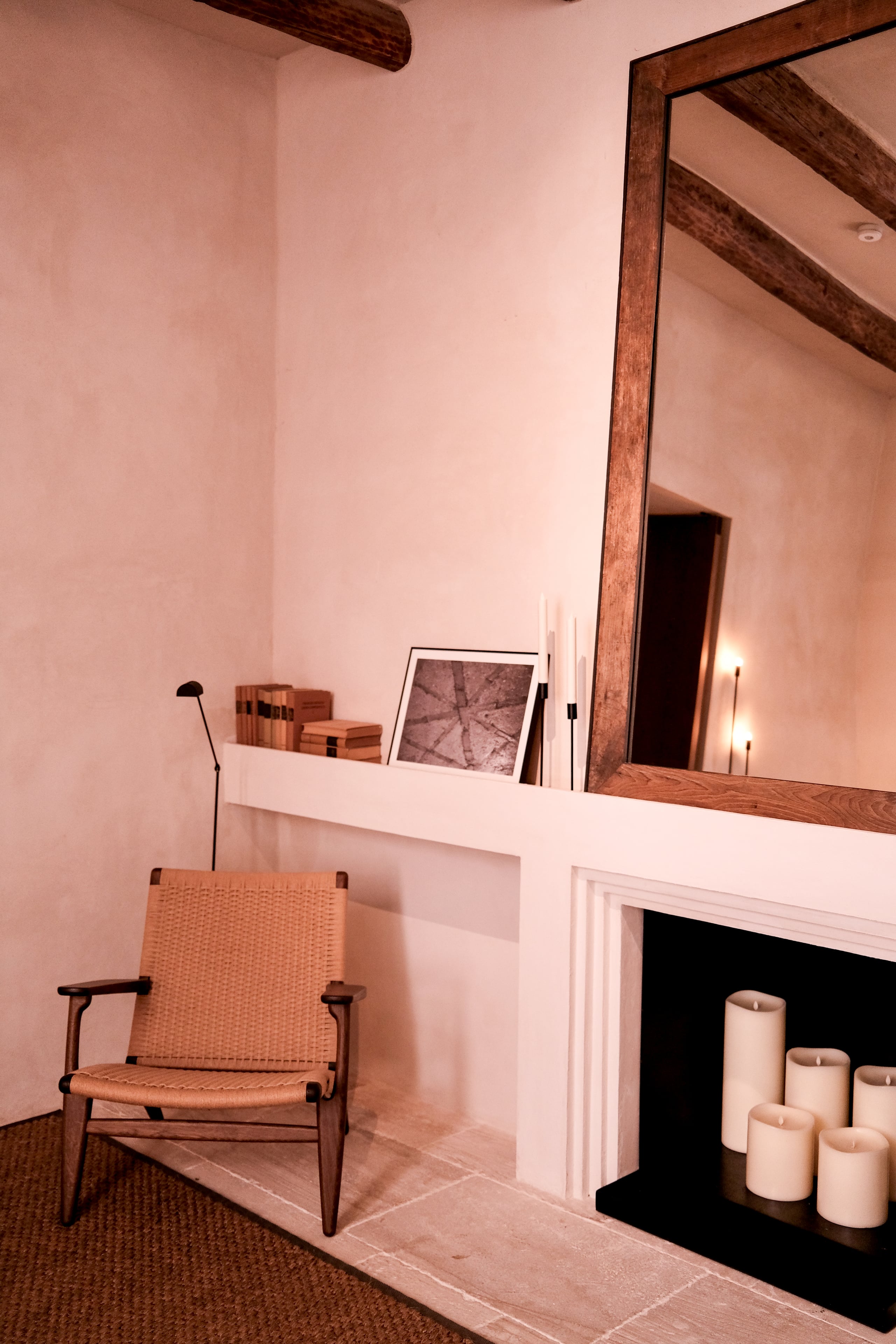 Can Ferrereta, Balearic Islands — Minimalist pink room with wooden-framed mirror, woven chair, fireplace with candles, exposed beams, and warm ambient lighting.