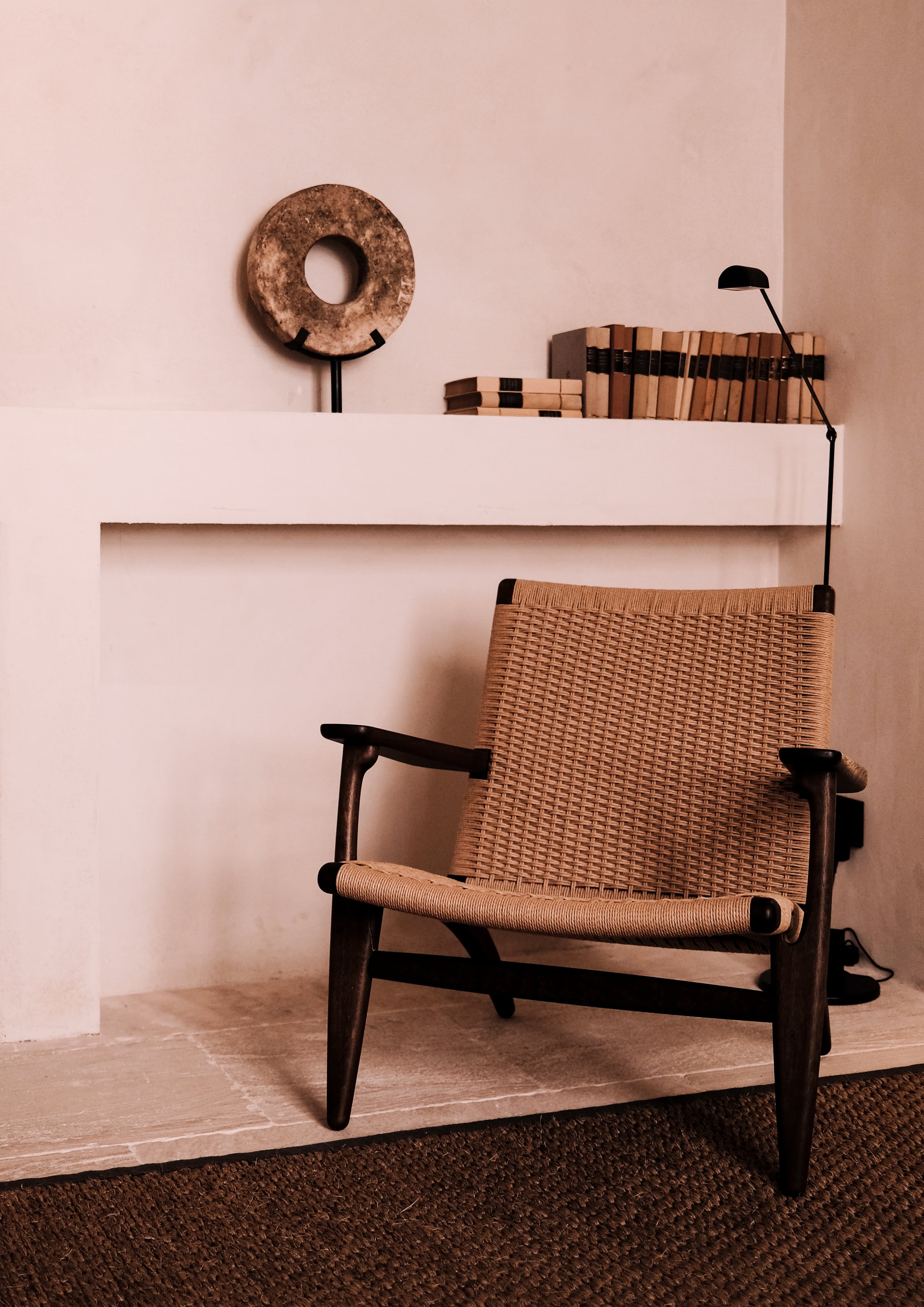 Can Ferrereta, Balearic Islands, Spain — Mid-century woven chair beside white shelving with stacked books, ceramic vessel, and black lamp in minimalist interior.
