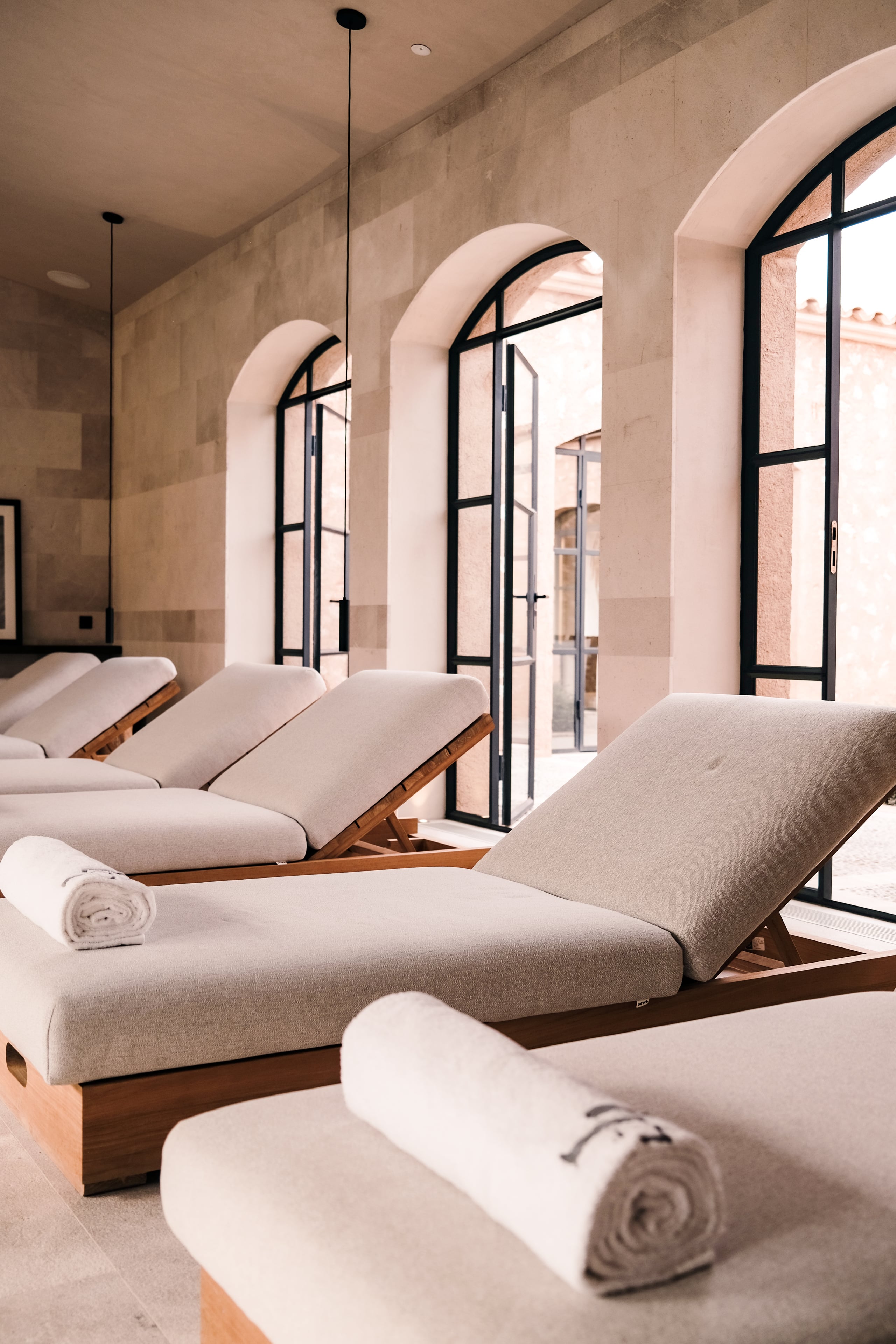 Can Ferrereta, Balearic Islands, Spain — Minimalist spa relaxation room with arched windows, wooden loungers upholstered in cream linen, rolled towels, concrete walls, and natural light.