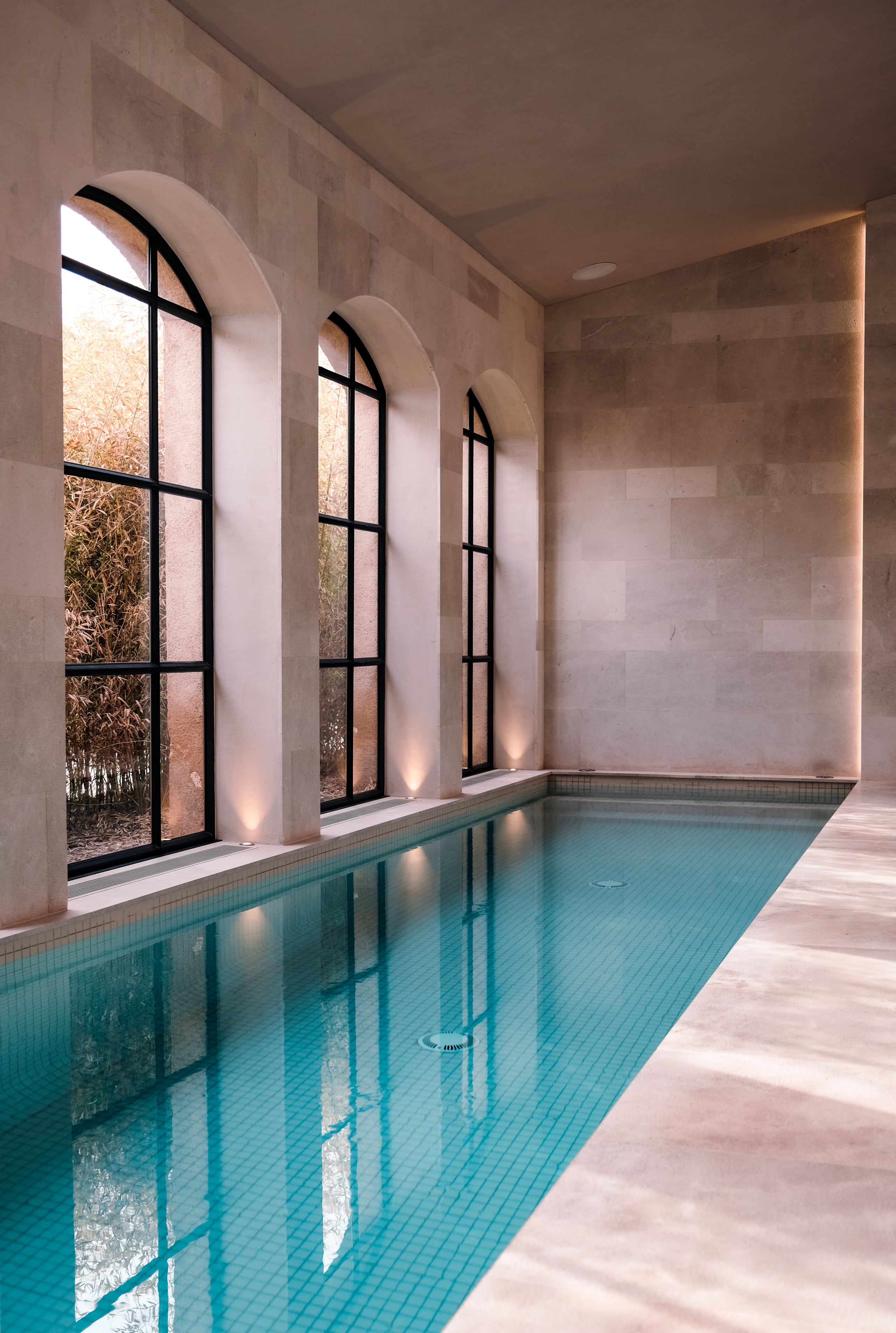Can Ferrereta, Balearic Islands, Spain — Indoor lap pool with arched windows, black-framed glass doors, warm stone walls, and terracotta tile flooring reflecting natural light.