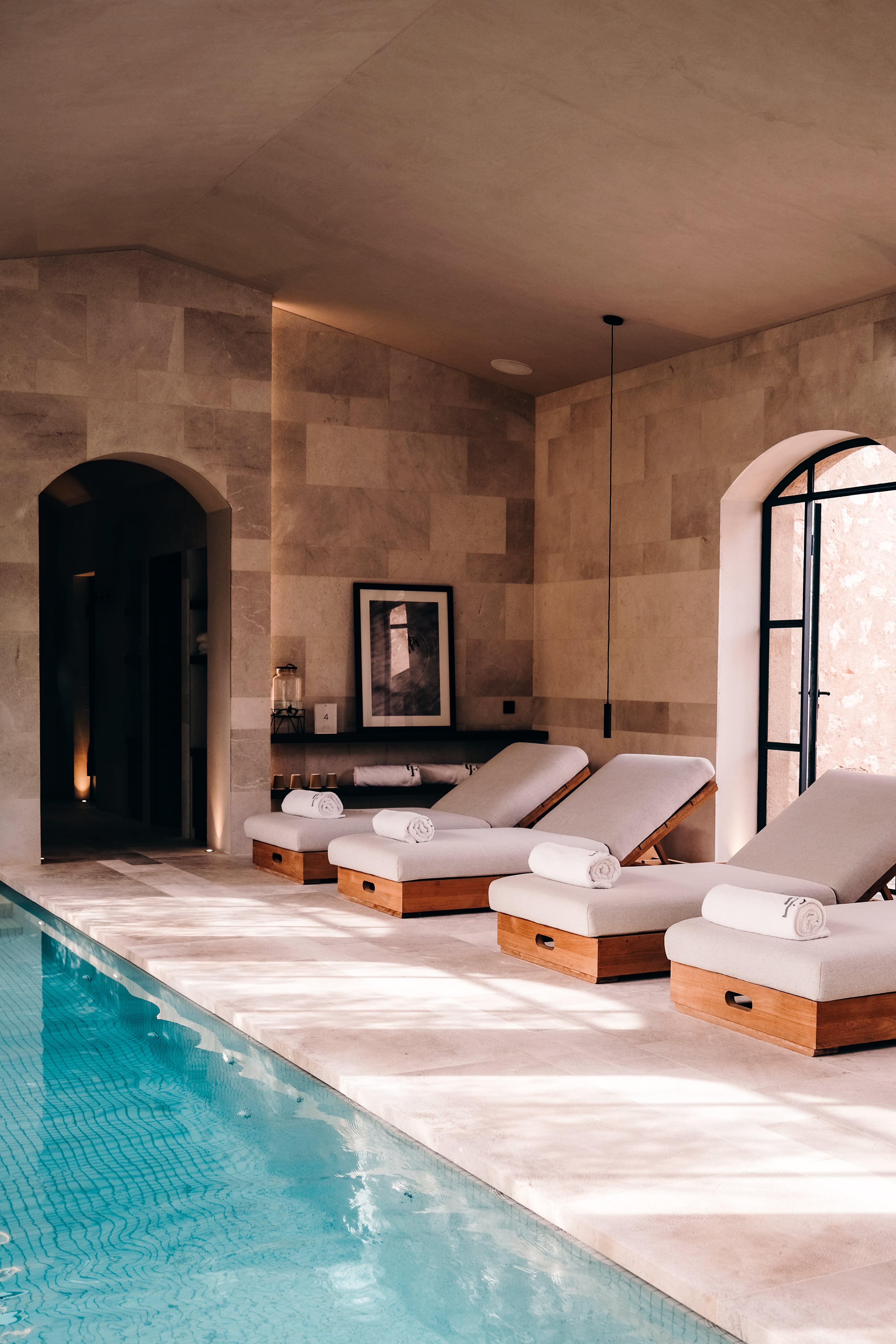 Can Ferrereta, Balearic Islands — Indoor pool with limestone walls, wooden loungers with cream cushions, arched doorways, and warm natural light filtering across terracotta stone flooring.