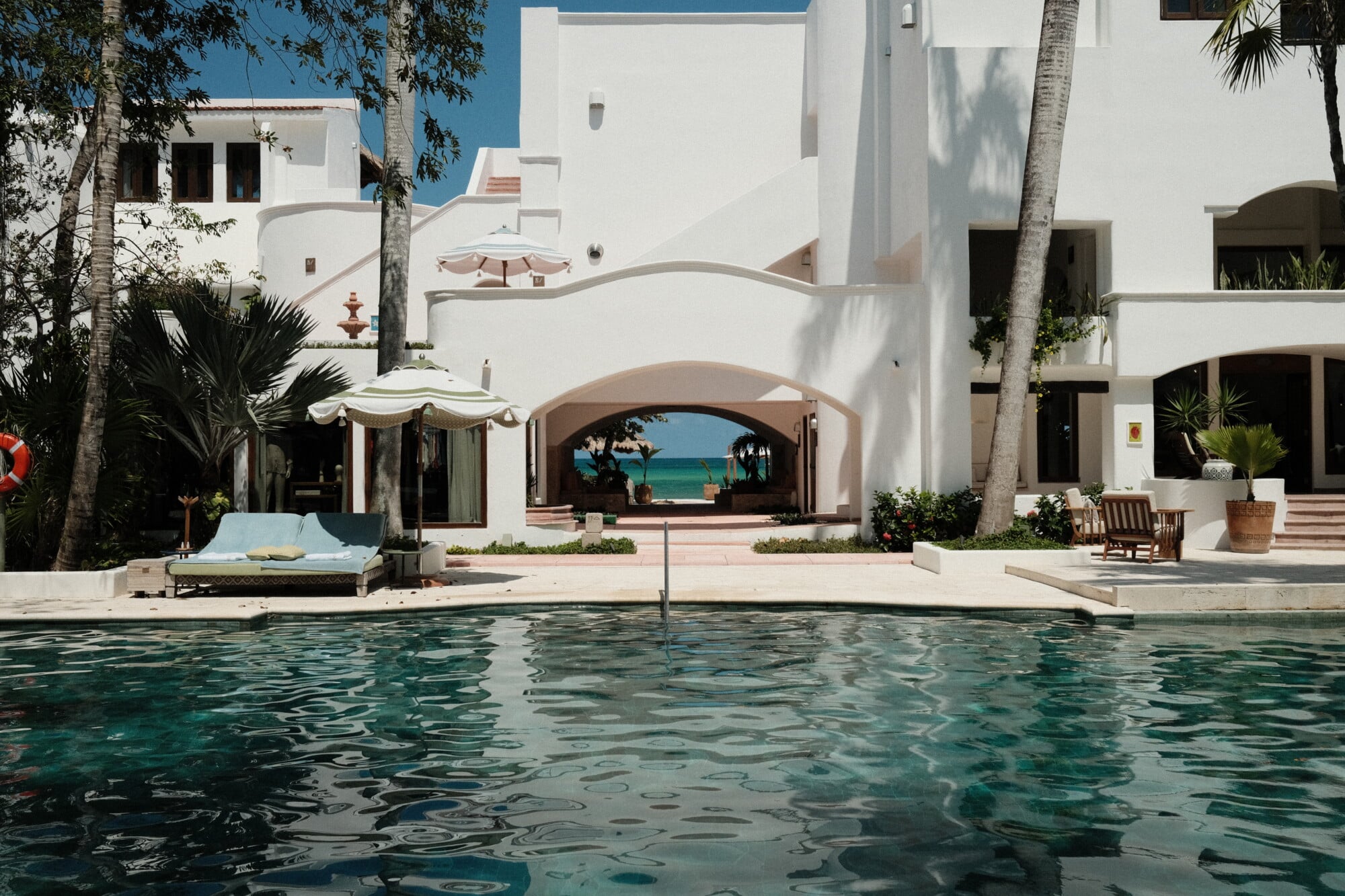 Maroma, A Belmond Hotel, Mexico — Modernist white courtyard with arched loggia framing turquoise sea view, palm trees, pool, and terracotta accents under clear sky.