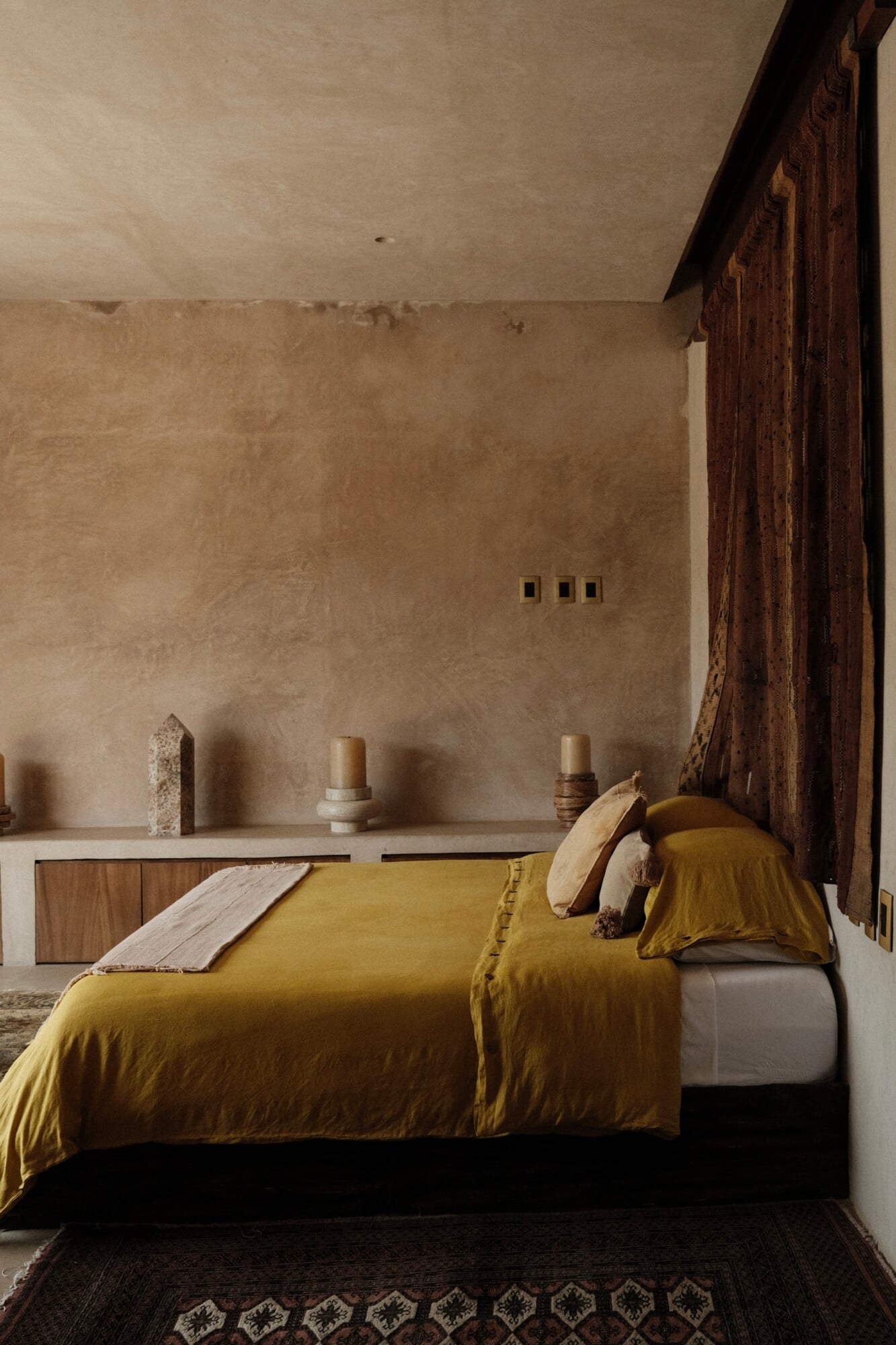 Casa Lumeria, Mexico — Minimalist bedroom with ochre linen bedding, plaster walls, carved wooden beam, ceramic vessels, and geometric patterned rug.