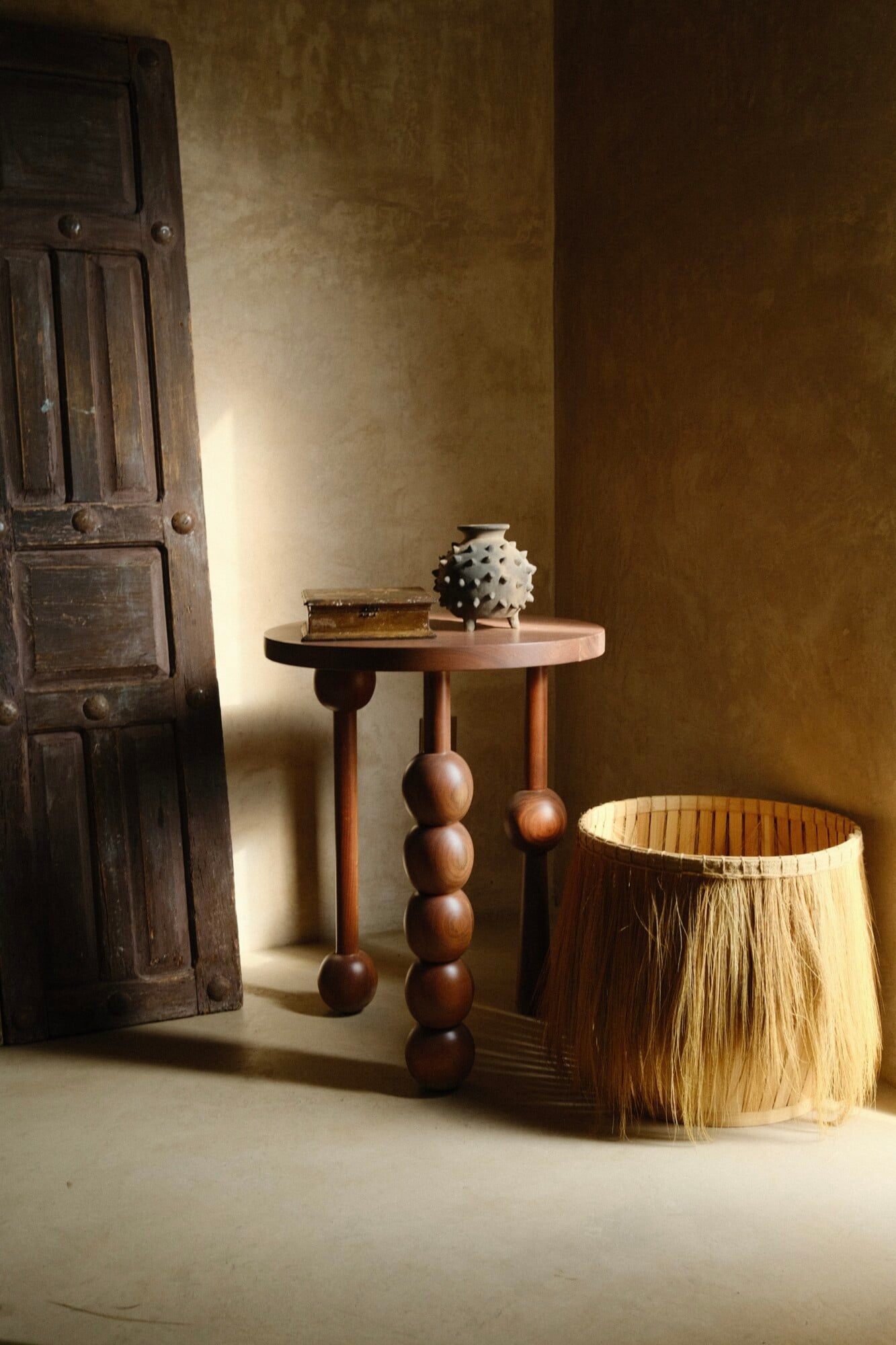 Casa Lumeria, Mexico — Wooden pedestal table with spherical legs beside woven basket, vintage door, and spiked ceramic vessel against ochre textured wall with natural light.
