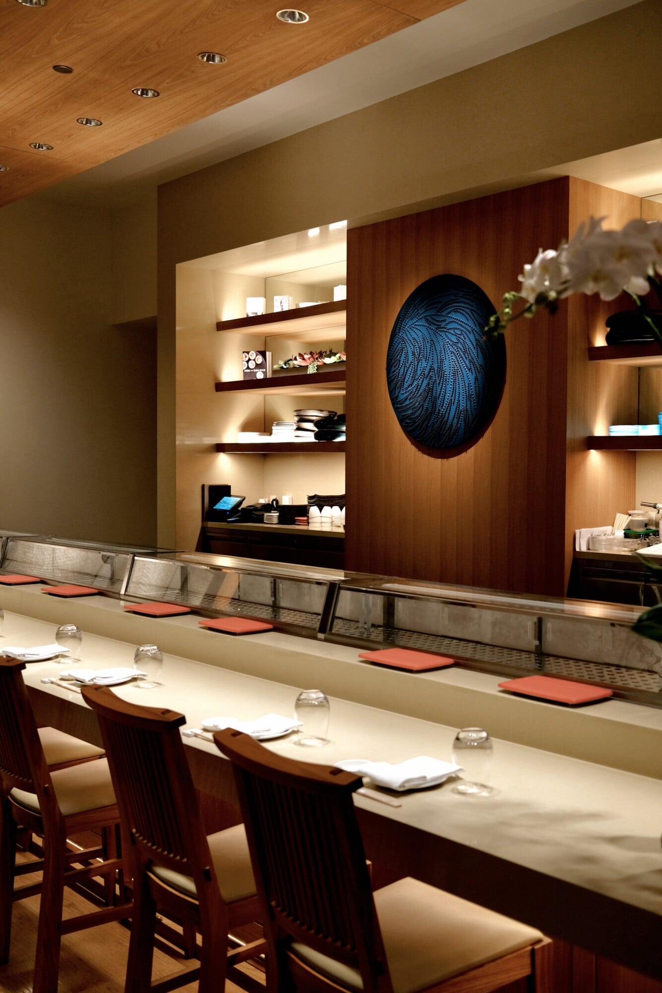Sensei by Nobu, North America — Sushi counter with wood paneling, illuminated shelving, blue circular artwork, and chef seating with coral cushions.