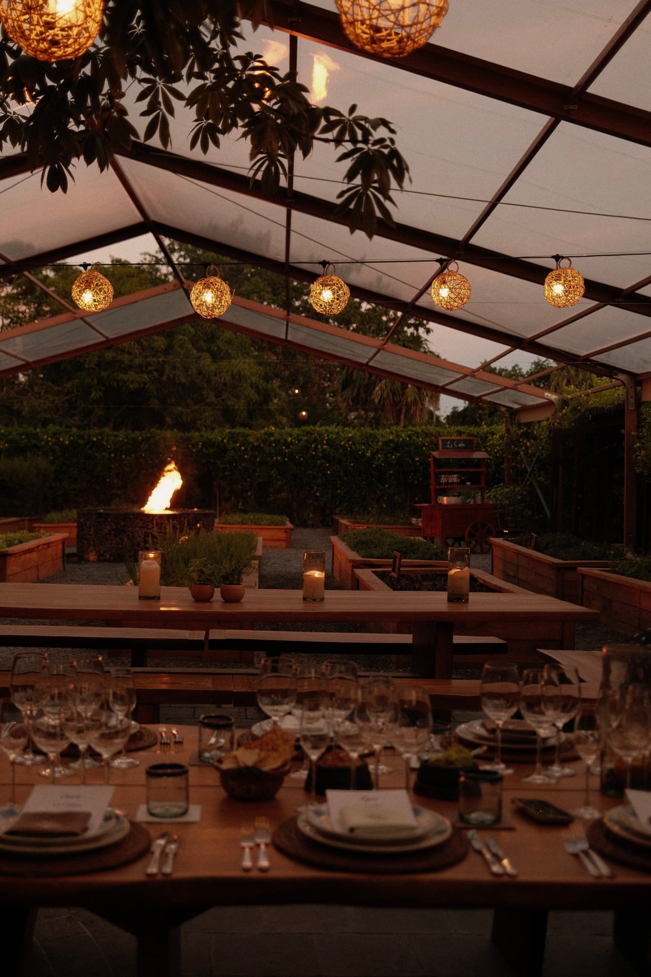 La Ceiba Garden & Kitchen, Mexico — Evening pergola dining with filigree pendant lights, candlelit table settings, fire pit glow, and manicured garden backdrop.