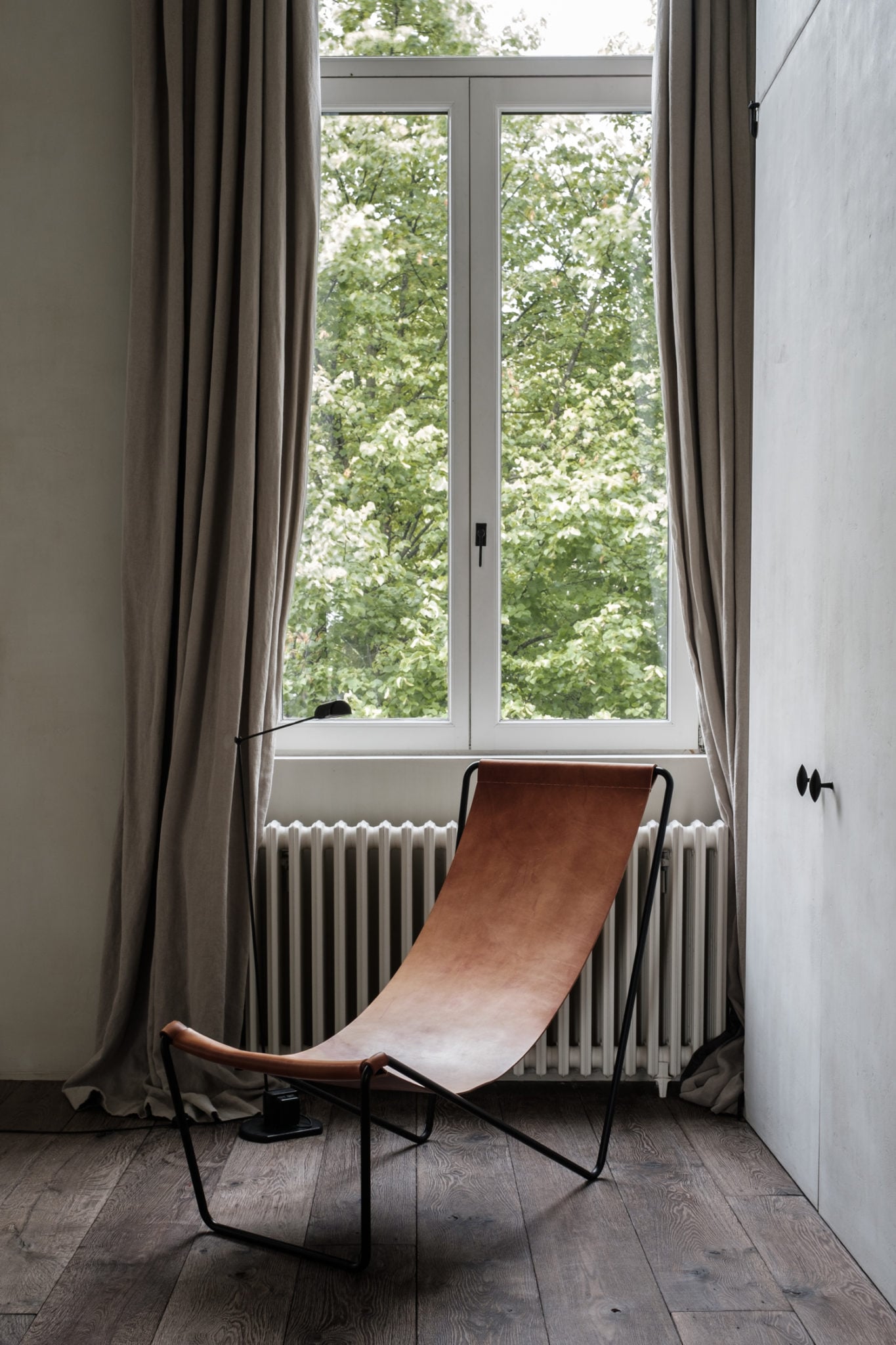 Ilse Cornelissens and Tim Van Geloven  | Graanmarkt 13 — Minimalist bedroom with cognac leather sling chair, tall windows overlooking flowering trees, radiator heating, natural light, contemporary Scandinavian design aesthetic.