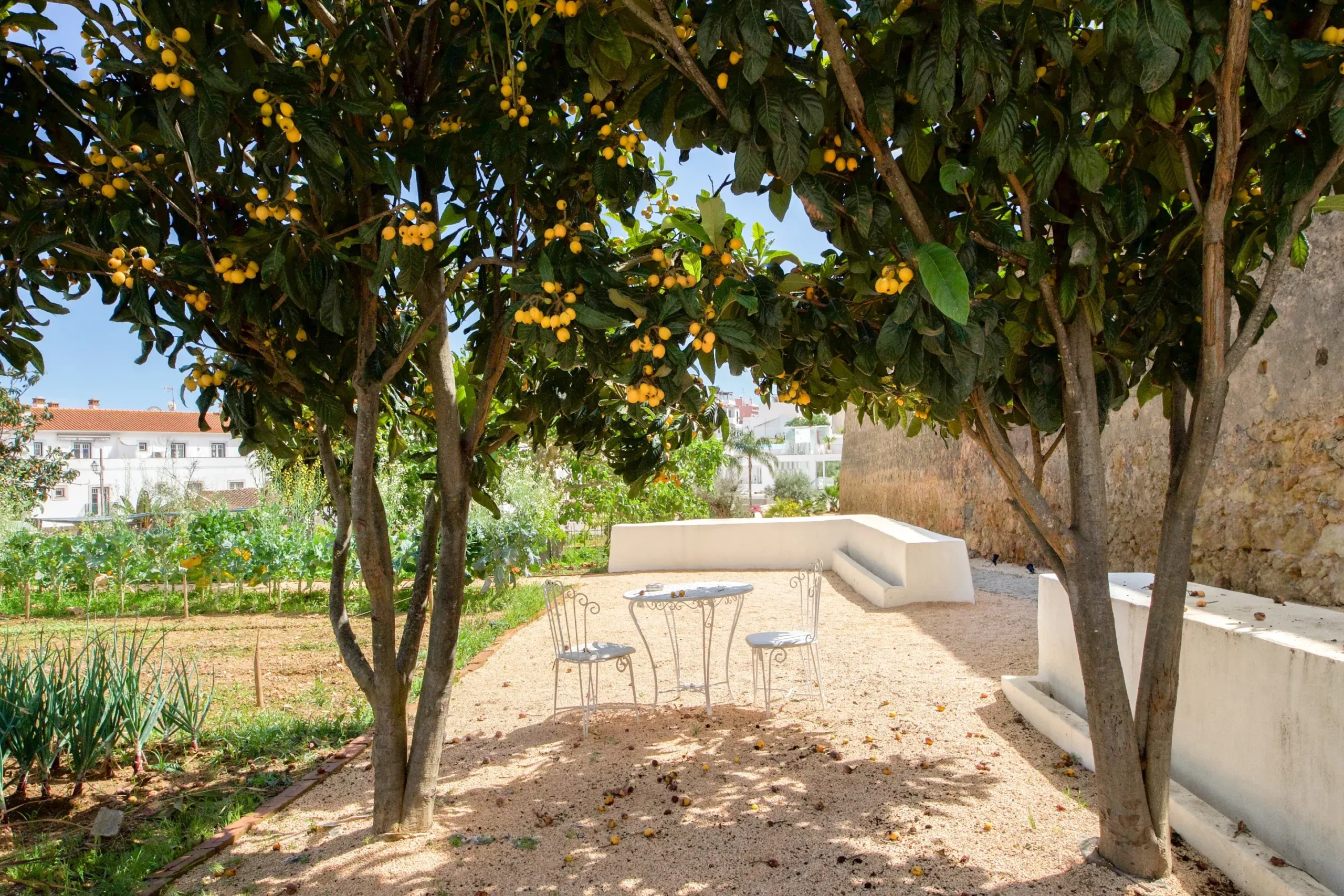 Casa Mãe, Europe — Shaded courtyard with yellow-fruited trees, wrought-iron seating, and stone walls overlooking terraced gardens and village rooftops.