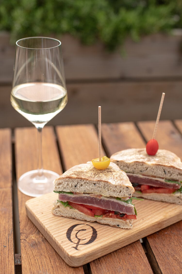 Bar Eira, Europe — Gourmet sandwiches with cured ham and fresh greens on wooden board beside crisp white wine, sunlit outdoor dining setting.