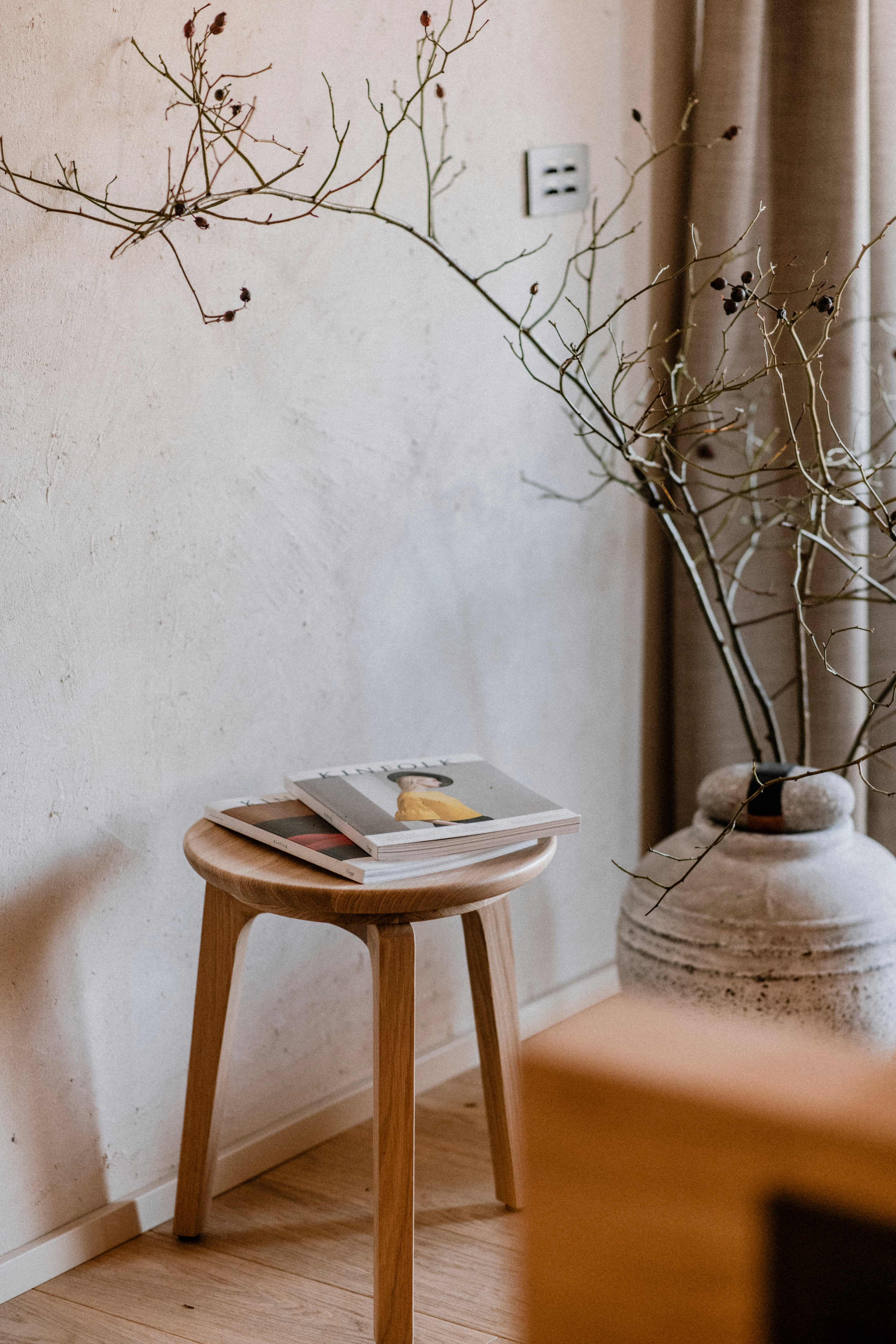 Casa di Langa, Europe — Minimalist wooden stool with stacked magazines beside dried botanicals in ceramic vase, natural light, neutral interior.
