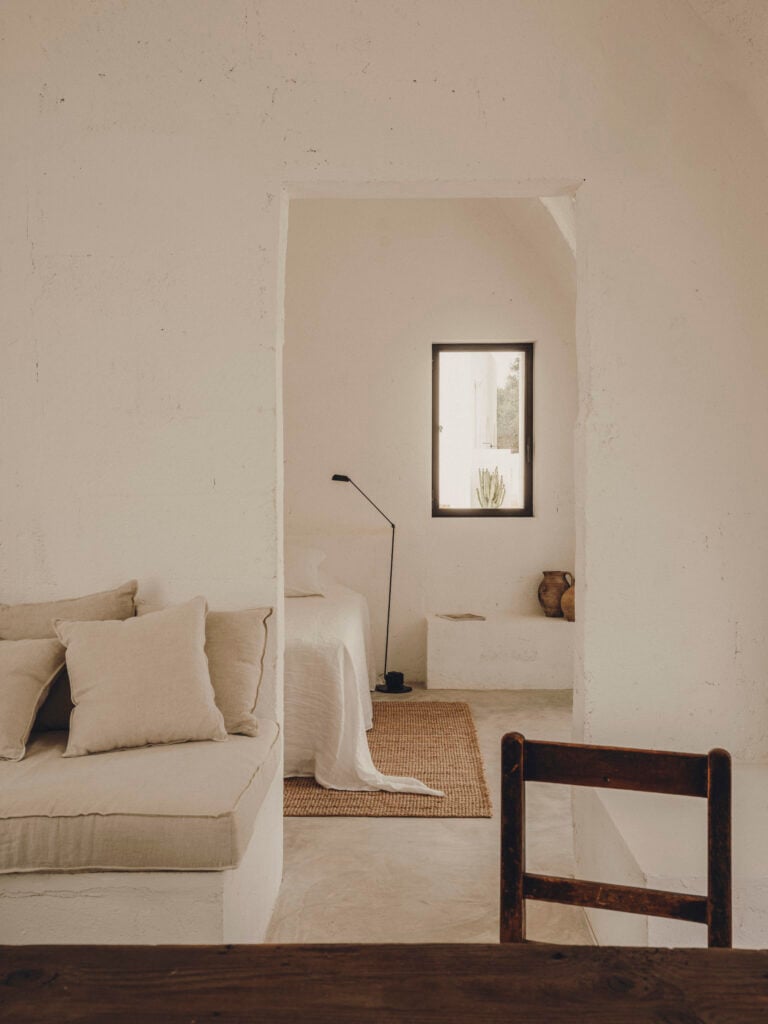 Borgo Gallana, Europe — Minimalist villa bedroom with cream linen furnishings, whitewashed walls, dark wood chair, and framed window overlooking landscape.