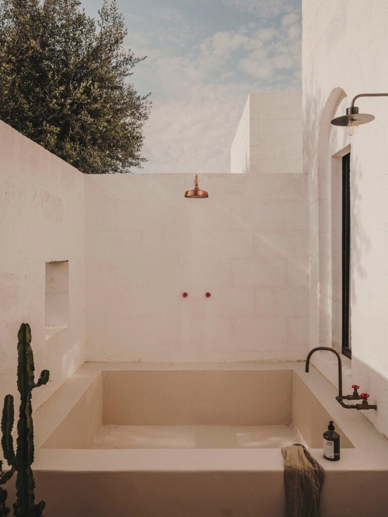 Borgo Gallana, Europe — Minimalist outdoor bathtub with copper shower fixtures, white plaster walls, and olive tree framed against sky.