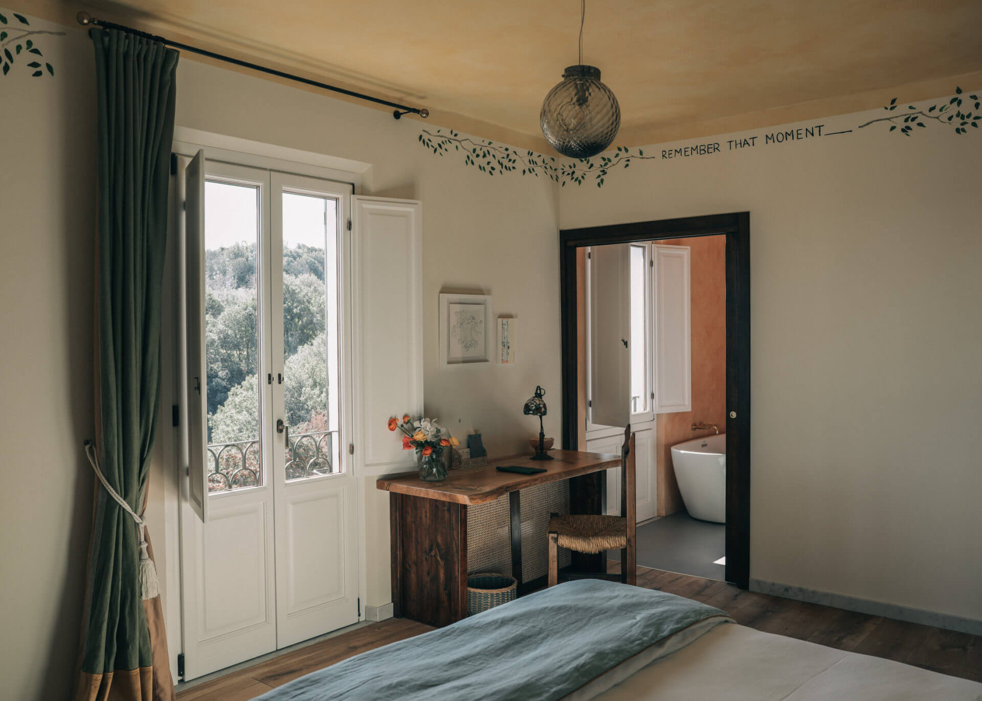 Villa Lena, Europe — Sunlit bedroom with wooden desk, open doors to balcony, modern bathroom, sage curtains, minimalist décor, and mountain views.