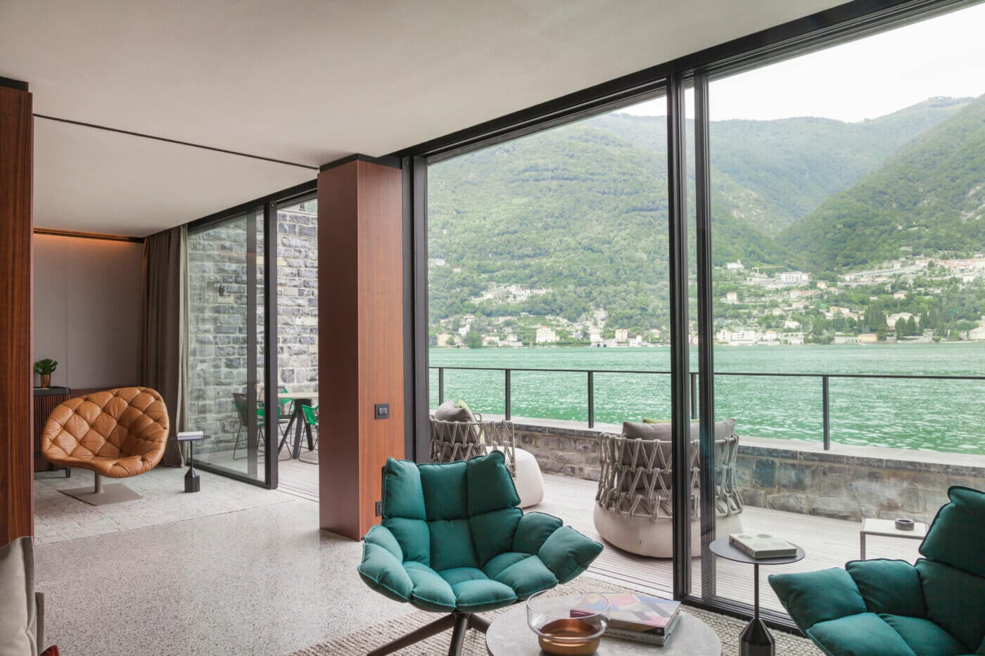 il Sereno Lago di Como, Europe — Modern lakeside suite with teal quilted chairs, cognac leather seating, floor-to-ceiling windows overlooking Alpine mountains and emerald water.