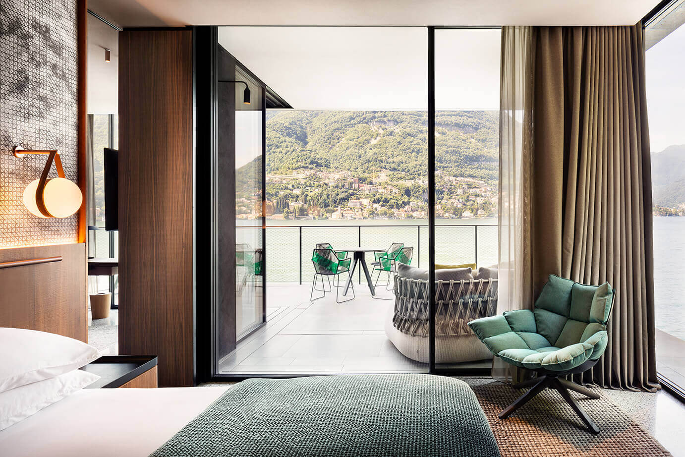 il Sereno Lago di Como, Italy — Modern lakeside bedroom with floor-to-ceiling windows overlooking Alpine mountains, walnut wood paneling, green accent chair, minimalist furnishings, and private terrace with dining setup.