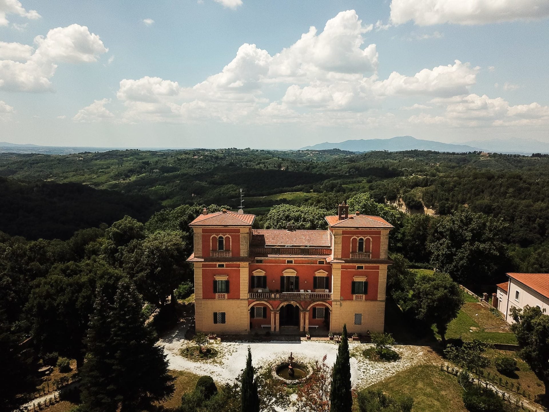 Villa Lena, Tuscany, Italy — Terracotta Renaissance villa with twin domed pavilions overlooking forested valleys and distant mountains under summer sky.