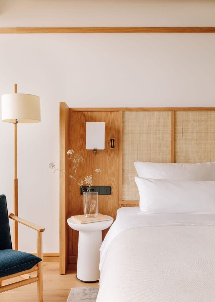 Aethos Ericeira, Portugal — Minimalist bedroom with natural wood headboard, crisp white linens, rattan details, and sculptural pedestal side table with dried flowers.
