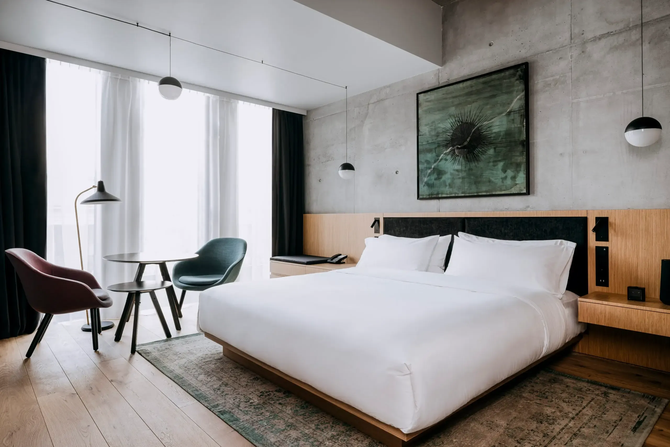Nobu Warsaw, Poland — Modern hotel bedroom with concrete walls, wooden bed frame, white linens, suspended pendant lights, and contemporary furnishings overlooking city views.
