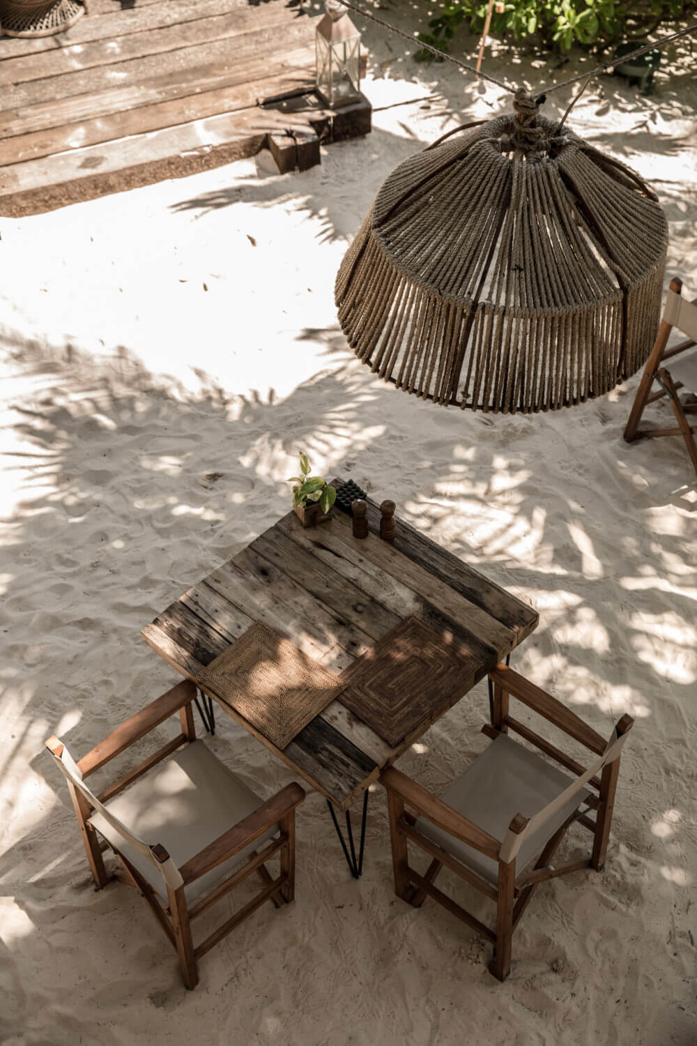 Be Tulum, Mexico — Wooden table and chairs on white sand beach with woven pendant umbrella and dappled shade from palm fronds.