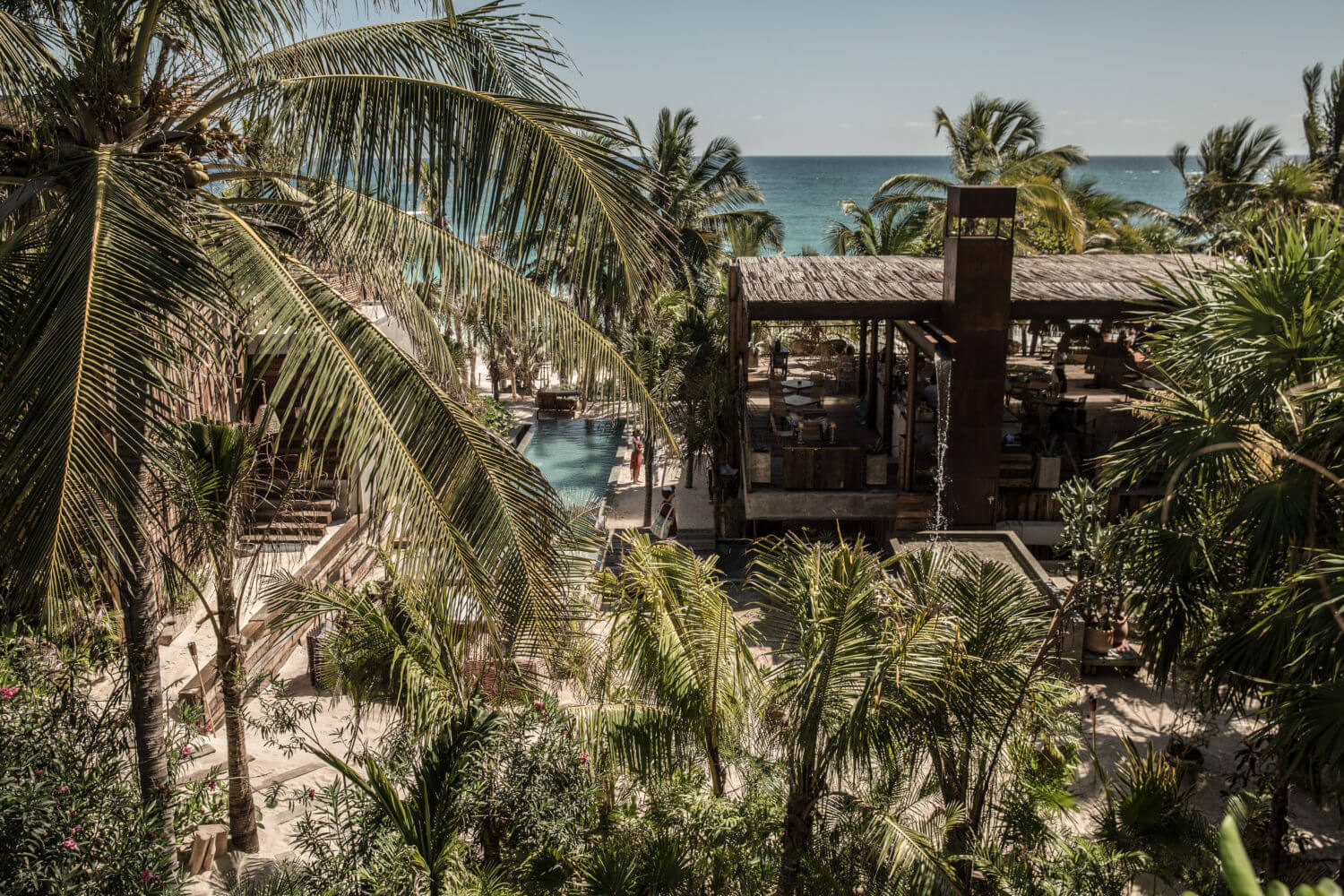 Be Tulum, Mexico — Palm-framed beachfront resort with dark wooden architecture, turquoise pool, and Caribbean Sea view under clear sky.