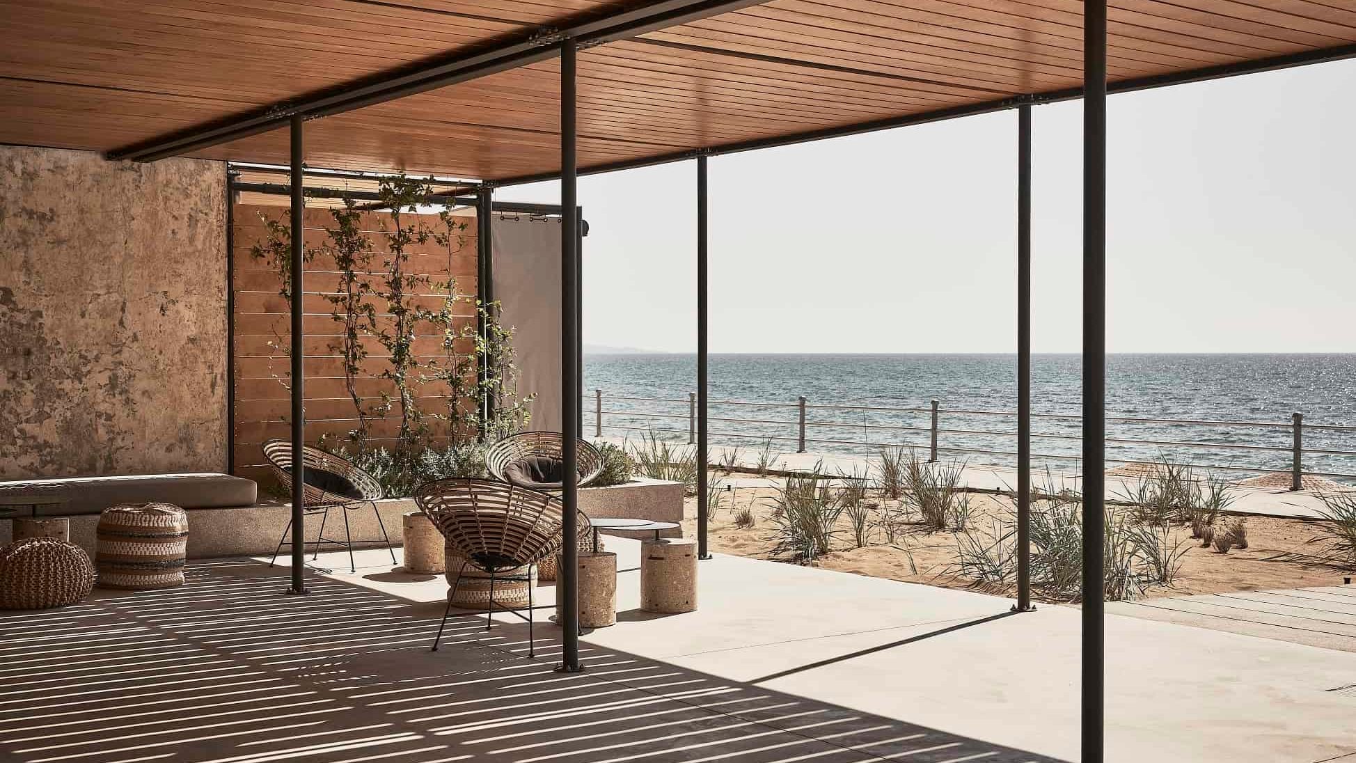 Dexamenes Seaside Hotel, Europe — Modern beachfront pavilion with wooden pergola, woven seating, and floor-to-ceiling glazing overlooking sand dunes and sea.