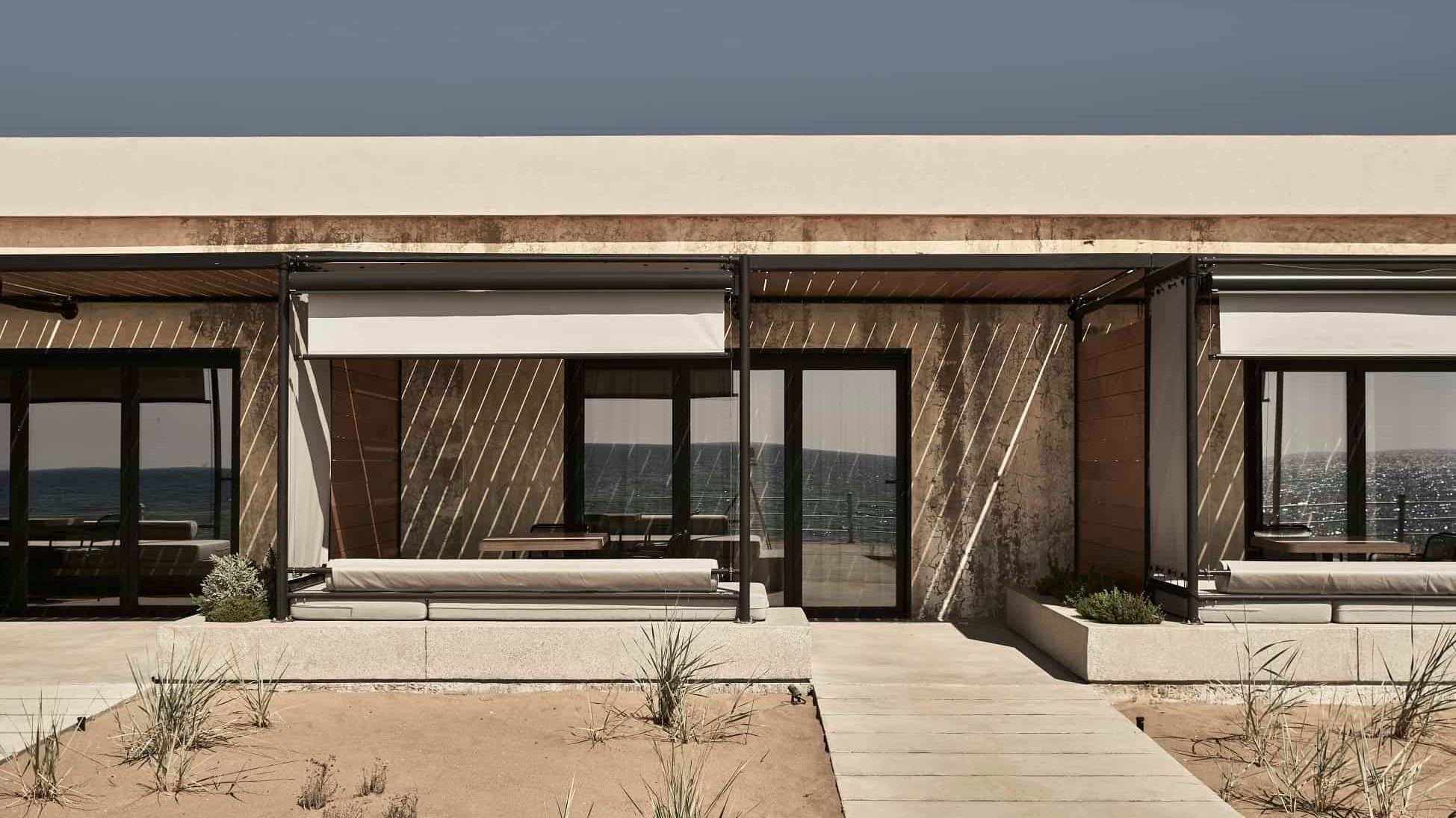 Dexamenes Seaside Hotel, Europe — Modern beachfront pavilion with slatted wood screens, concrete terraces, and floor-to-ceiling glazing overlooking calm sea.