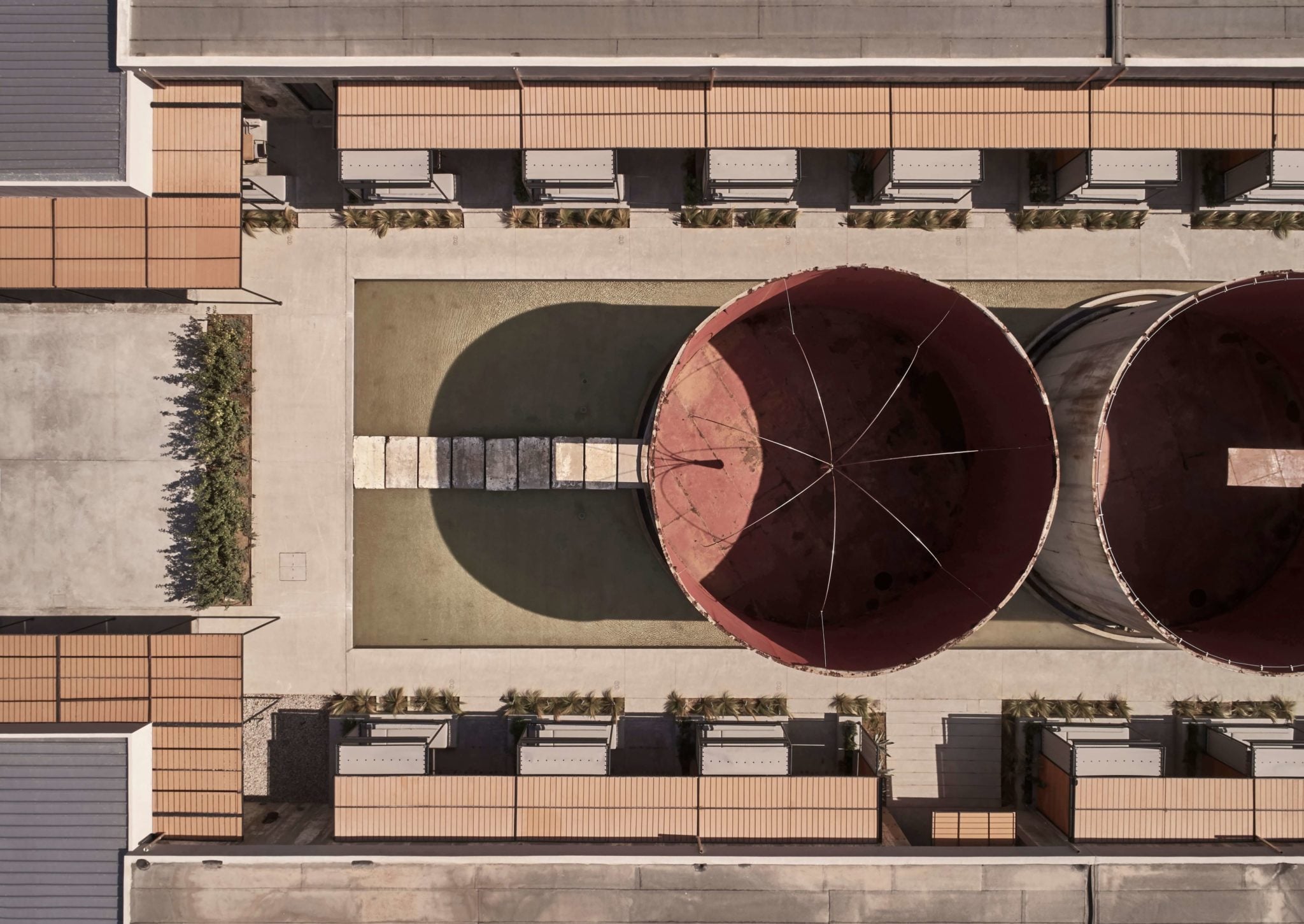 Nikos Karaflos | Dexamenes Seaside Hotel — Aerial view of industrial storage tanks positioned in modernist courtyard with terracotta brick residential buildings and manicured plantings, Mediterranean coastal architecture.