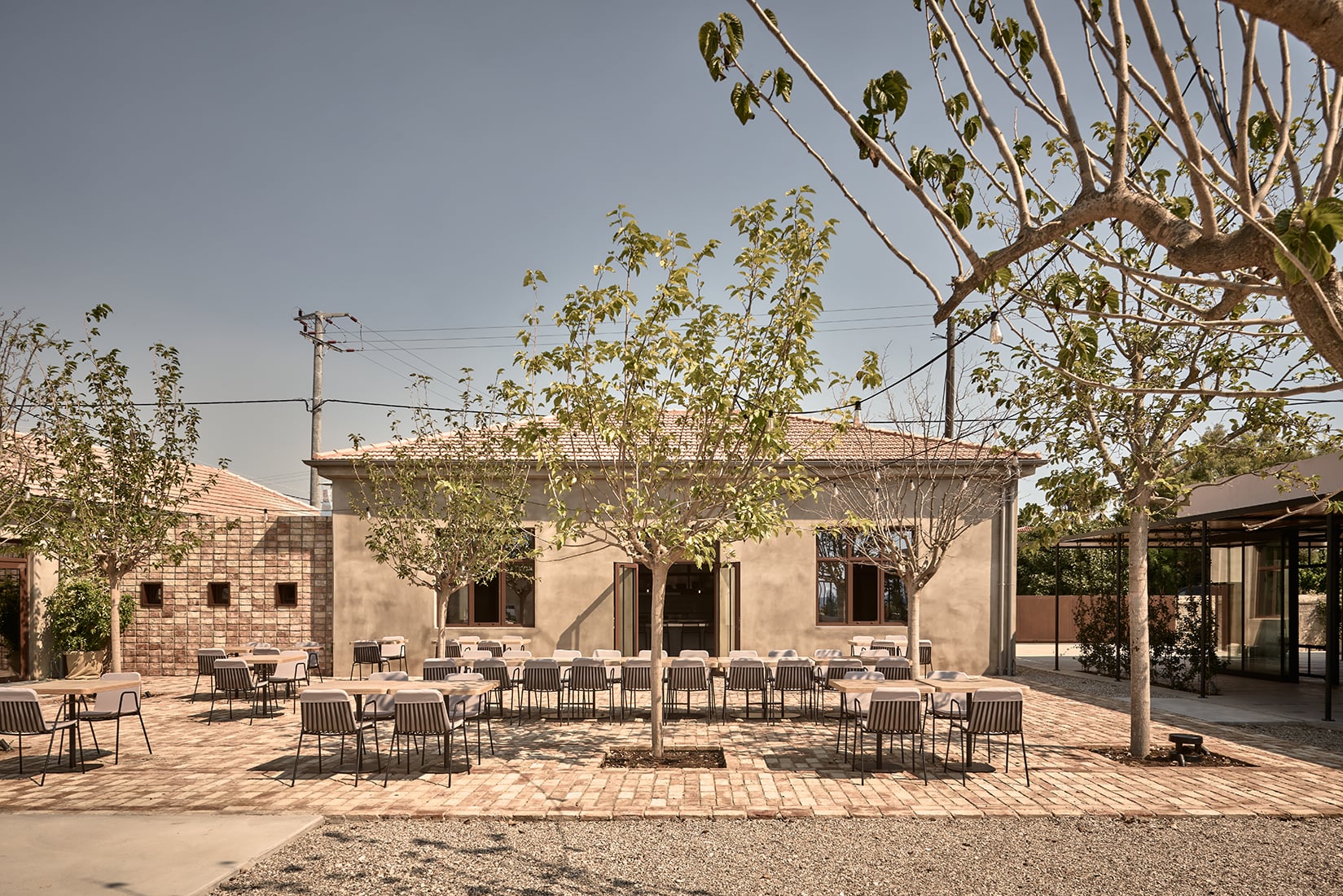 Dexamenes Seaside Hotel, Europe — Courtyard with terracotta paving, modern rope chairs, mature trees, and minimalist stone architecture under clear sky.