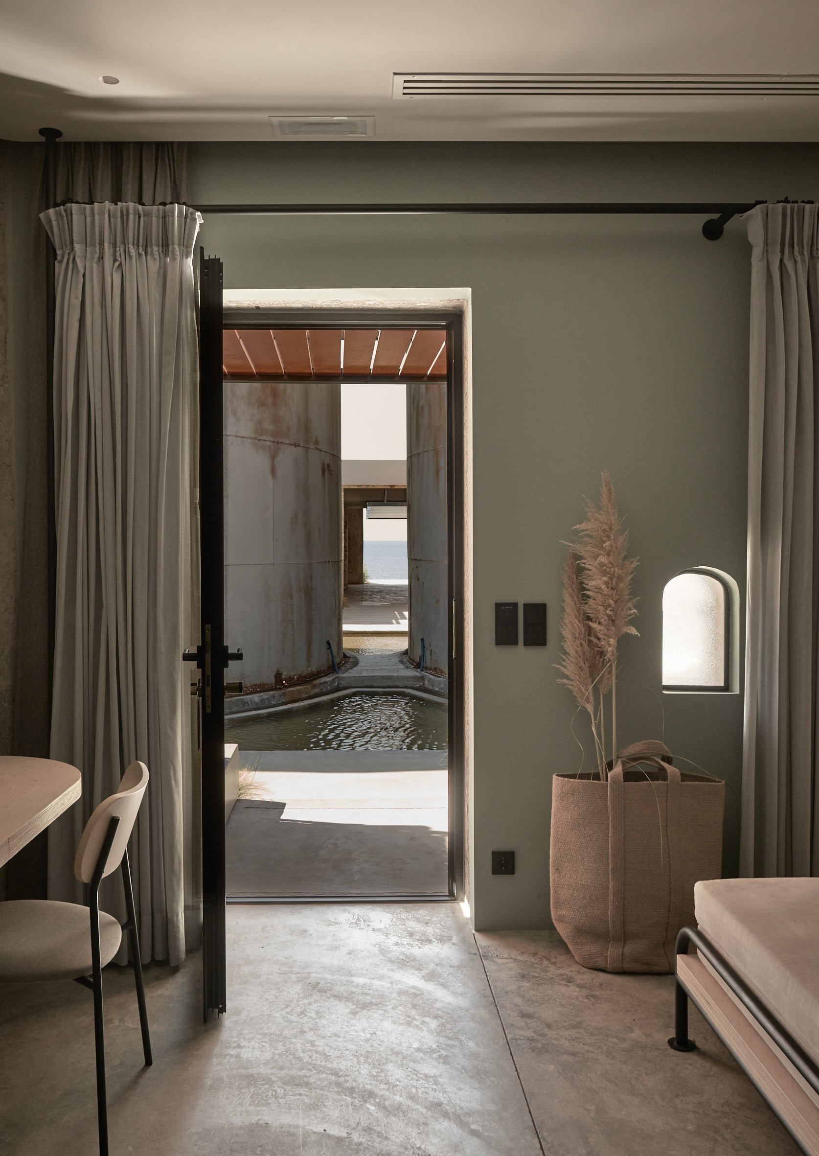 Dexamenes Seaside Hotel, Europe — Minimalist bedroom with concrete floors, sage walls, linen curtains, opening to spa with soaking tub and sea views.