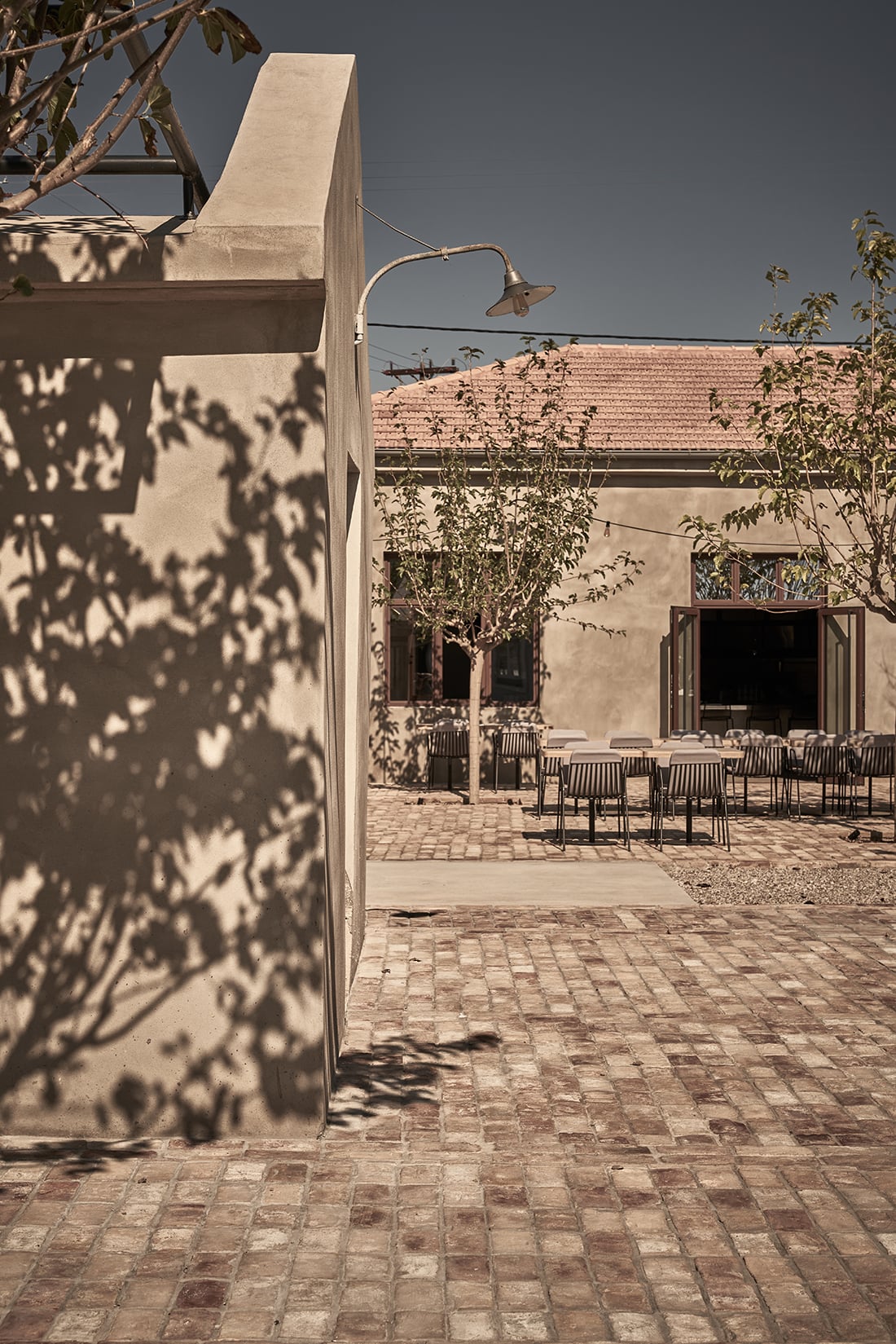 Dexamenes Seaside Hotel, Europe — Minimalist courtyard with terracotta brick paving, concrete walls casting leaf shadows, dining chairs arranged under a pitched roof structure, warm Mediterranean light.