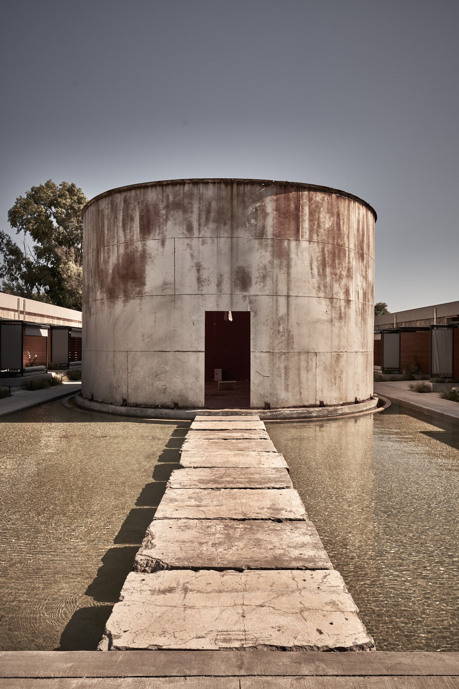 Dexamenes Seaside Hotel, Europe — Weathered cylindrical concrete structure with rust staining, dark doorway, stepping stones, minimalist courtyard design under clear sky.