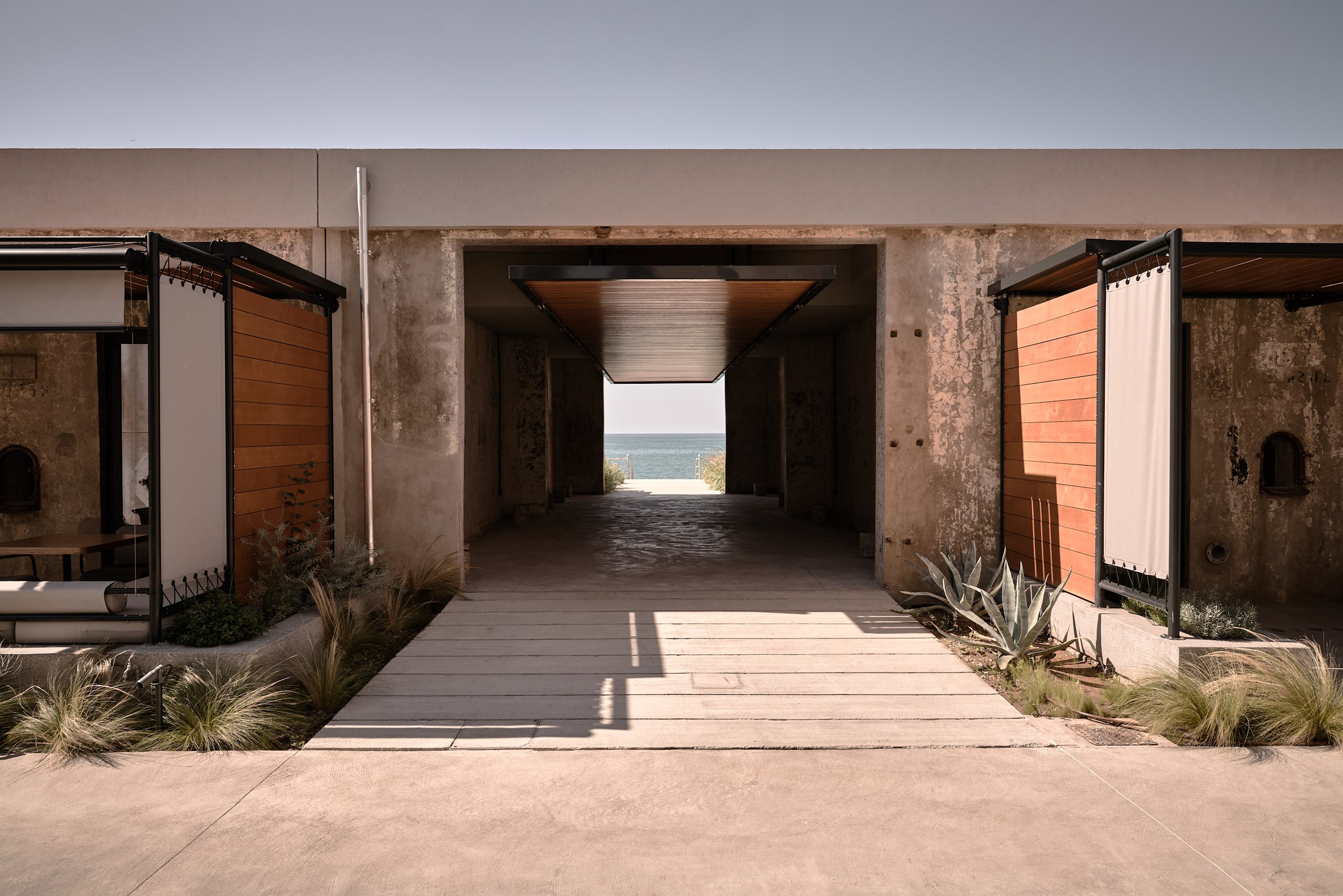 Dexamenes Seaside Hotel, Europe — Concrete corridor with wooden pergolas and terracotta shutters framing ocean view, minimalist coastal architecture.