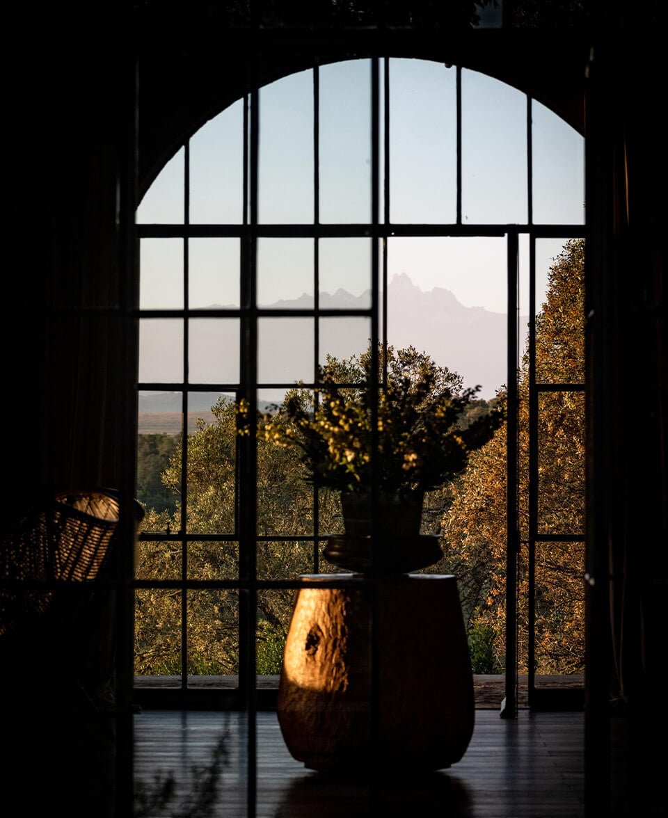 Arijiju Retreat, Africa — Arched window with gridded panes framing mountain vista, silhouetted potted plant catching golden afternoon light, luxury villa interior overlooking savanna landscape.