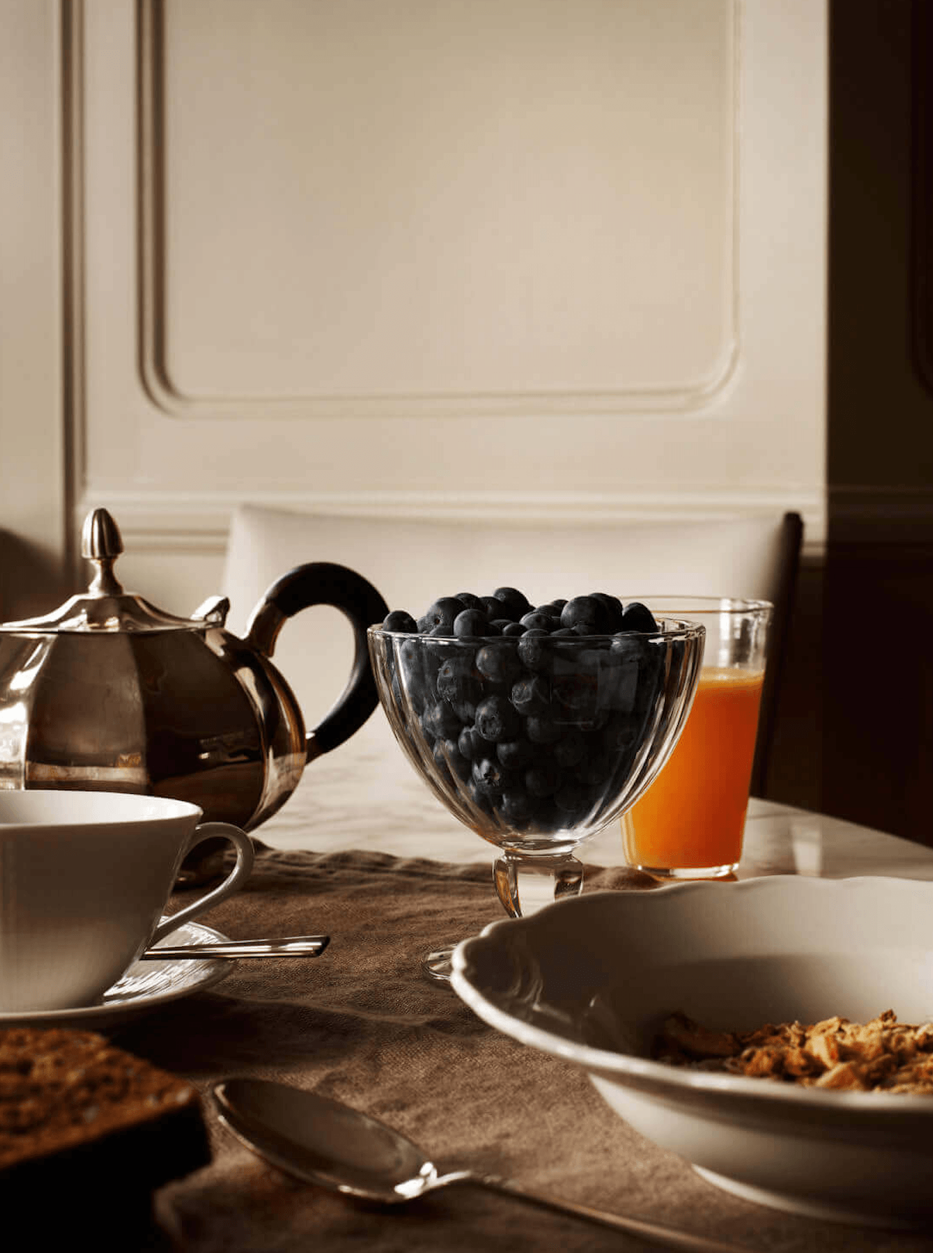 Ett Hem, Europe — Elegant breakfast table with blueberries in crystal bowl, silver teapot, amber juice, and natural morning light through paneled wall.