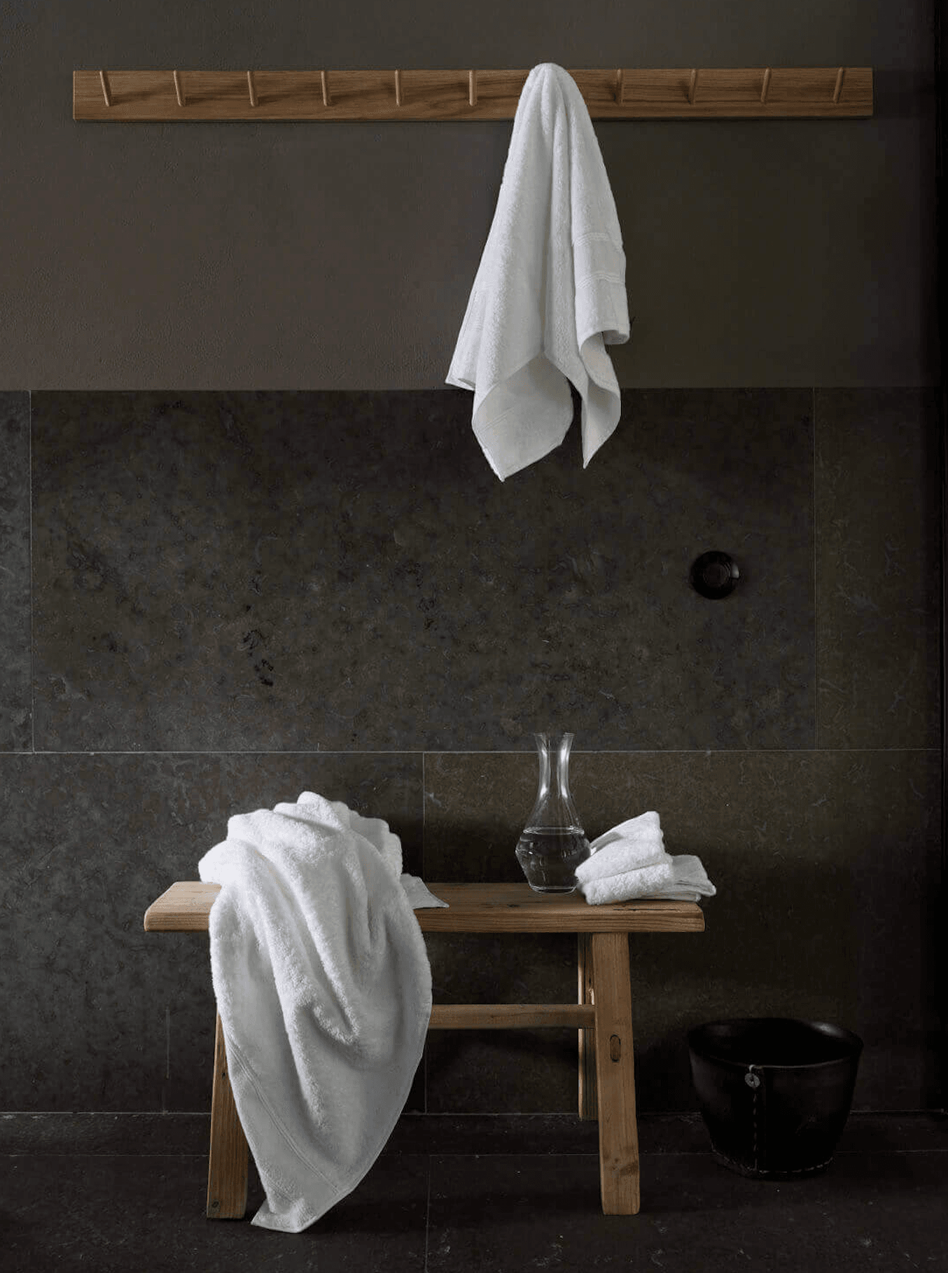 Ett Hem, Europe — Minimalist spa interior with dark stone walls, natural wood bench, white linens, glass vessel, and wooden peg rail.