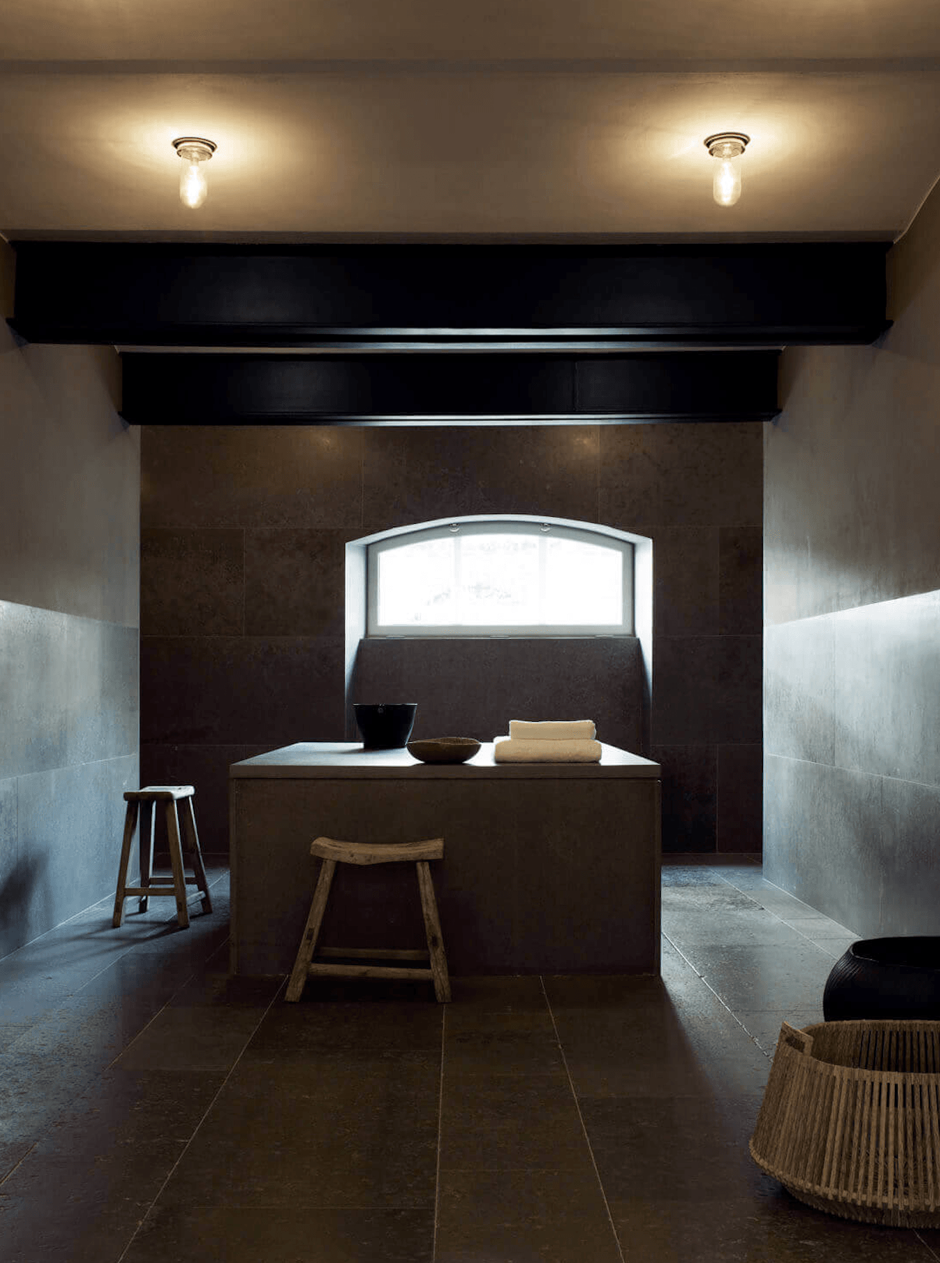 Ett Hem, Europe — Minimalist spa bathroom with dark tile walls, sunlit transom window, wooden stools, and natural fiber baskets creating serene Japanese-inspired wellness space.