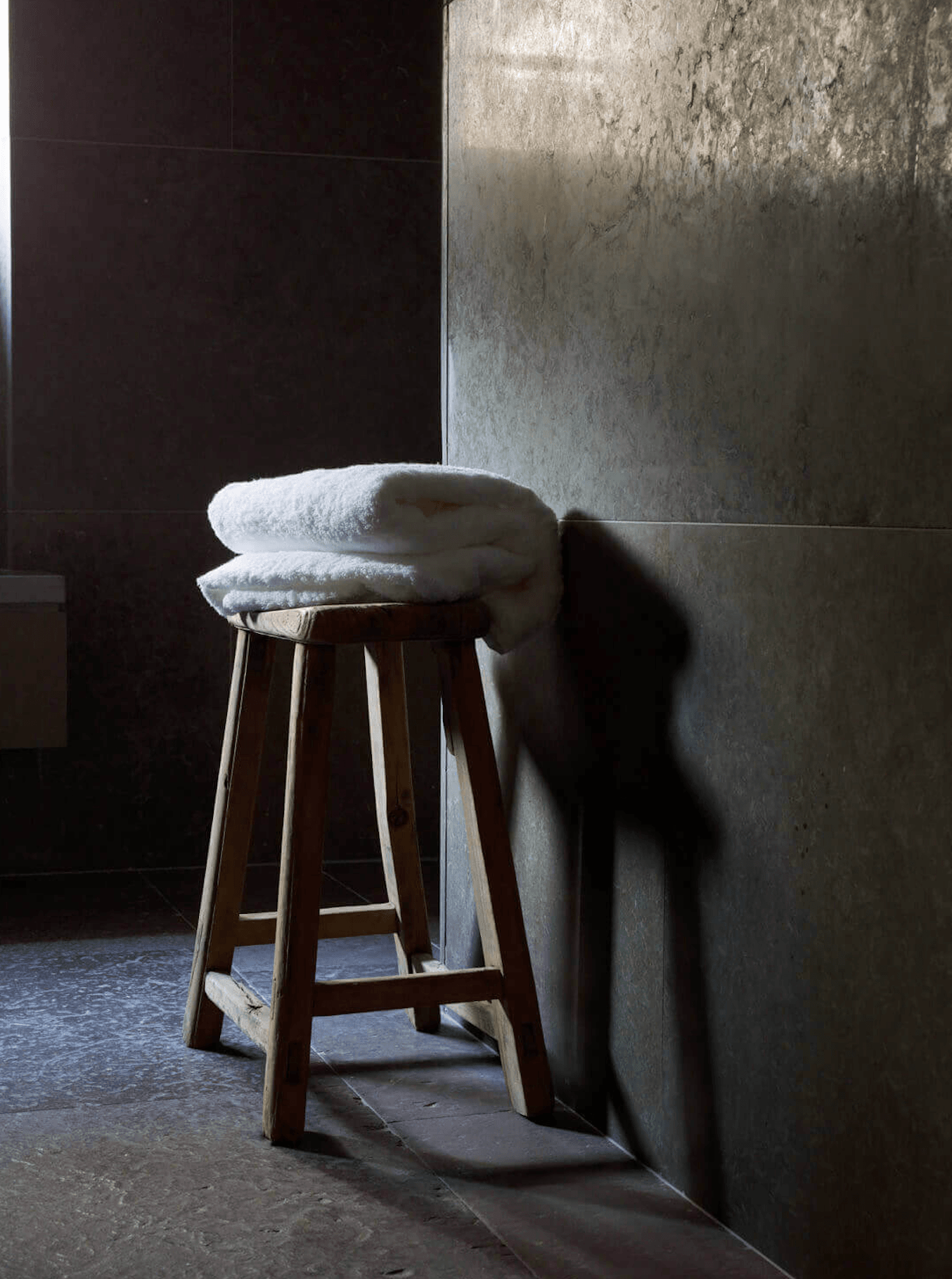 Ett Hem, Europe — Wooden stool with folded white towels against weathered concrete wall in minimalist bathroom with natural light and shadow.