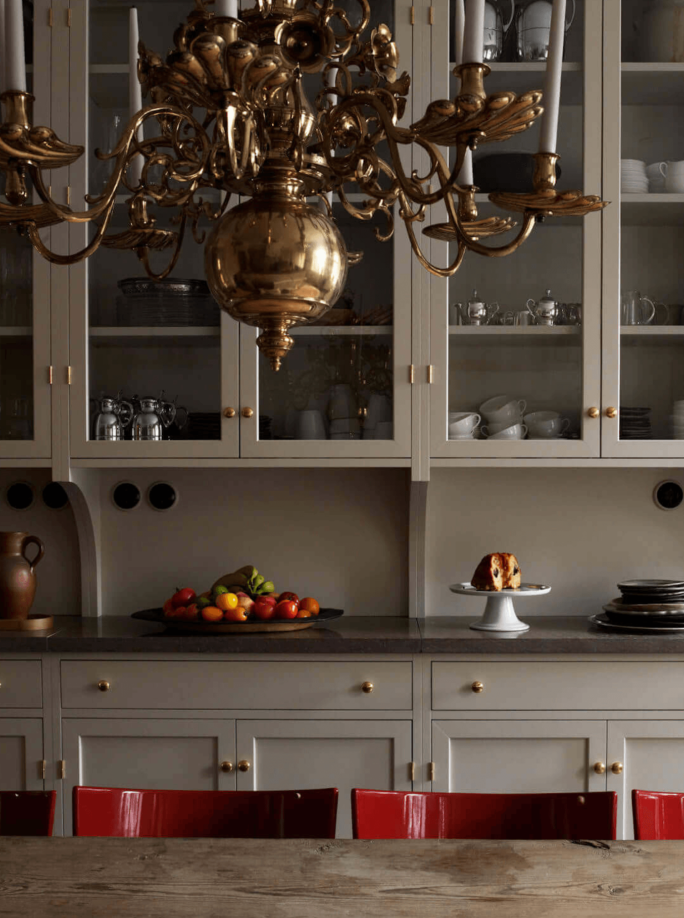 Ett Hem, Europe — Brass chandelier above cream cabinetry with glass doors, marble countertop, fresh fruit display, and burgundy dining chairs.