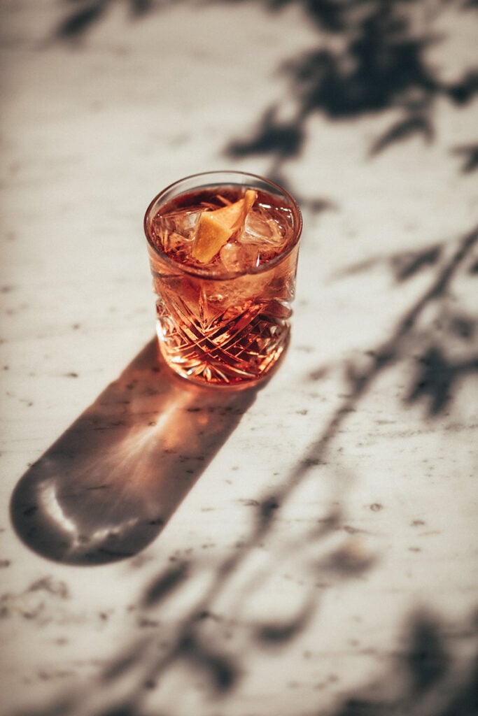 Epi 1959, Europe — Amber cocktail with ice and citrus peel casting shadow on speckled stone surface in natural sunlight.