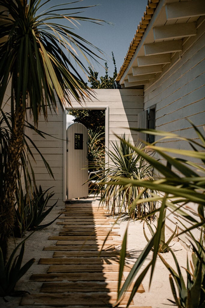 Epi 1959, Europe — Coastal pathway with wooden decking, white-rendered walls, palm fronds, and modernist architecture casting geometric shadows in bright sunlight.