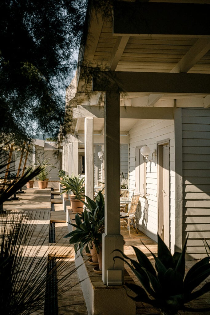 Epi 1959, Europe — Covered terrace with timber pergola, potted plants, and wicker seating overlooking whitewashed villa, dappled shade from mature trees.