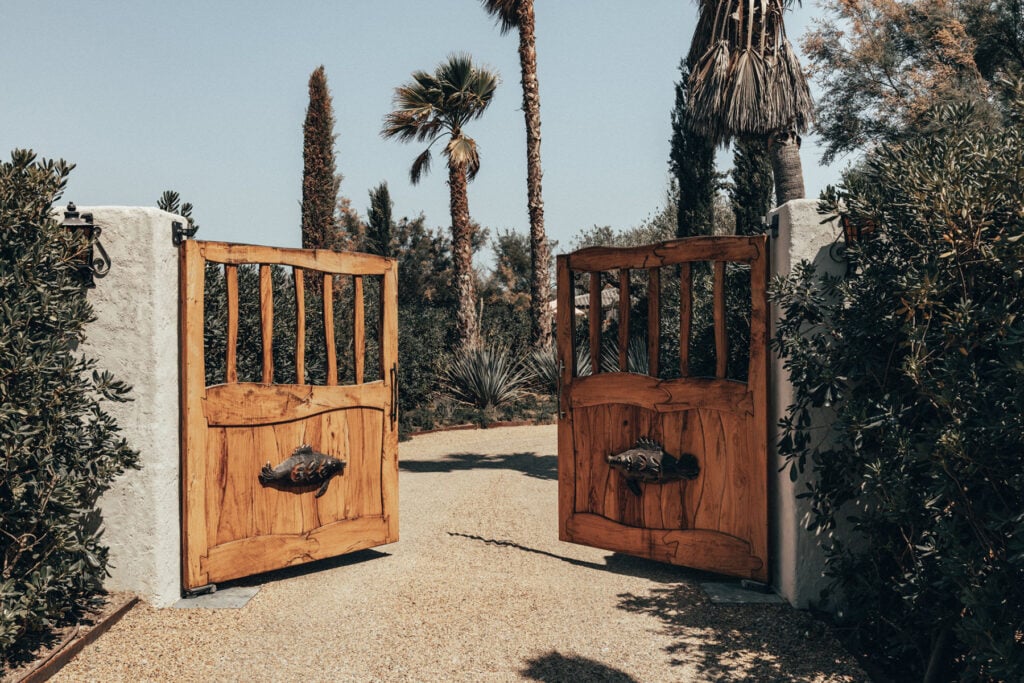 Epi 1959, Europe — Wooden estate gates with vertical paneling and iron hardware open onto palm-lined gravel driveway under clear sky.