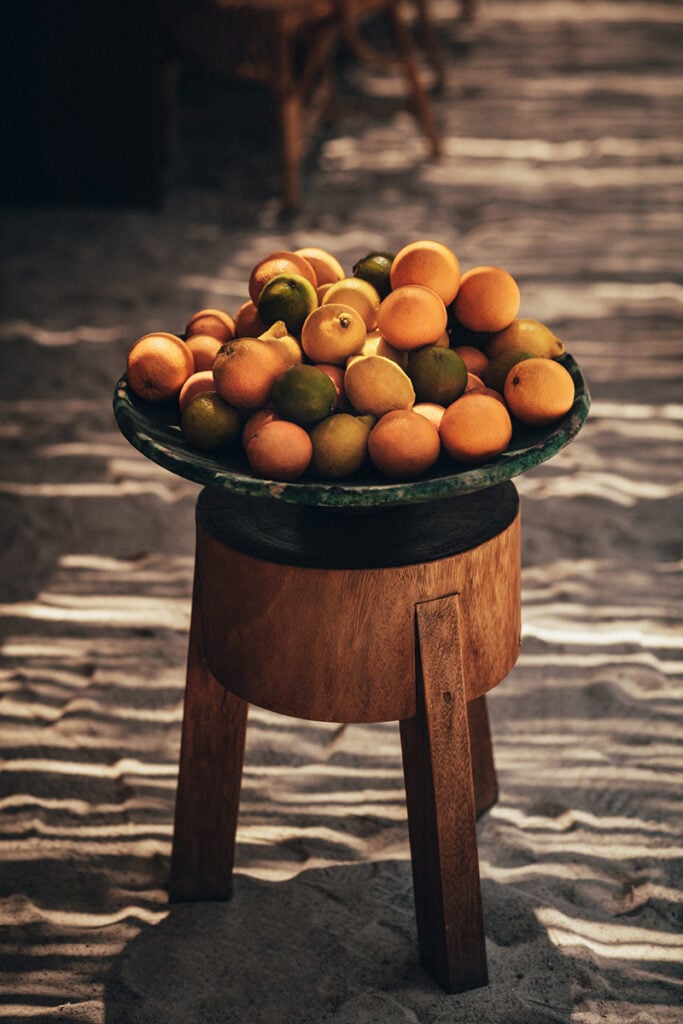 Epi 1959, Europe — Wooden pedestal table with weathered metal bowl displaying oranges and limes on sandy ground, warm golden light.