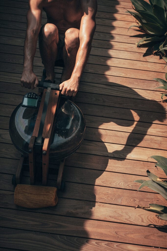 Epi 1959, Europe — Shirtless man using exercise equipment on wooden deck in dappled sunlight, minimalist fitness aesthetic.