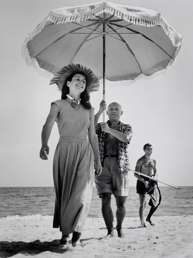 Epi 1959, Europe — Woman in linen dress and wide-brimmed hat walks beach with umbrella, vintage photograph, mid-century leisure aesthetic.