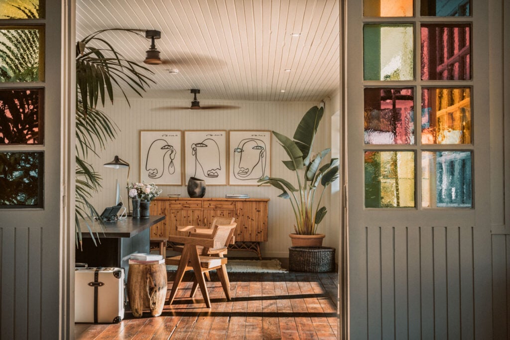 Epi 1959, Europe — Minimalist studio workspace with lined ceiling, line-drawn portraits, wooden furniture, potted plants, and colorful stained-glass windows casting warm light across terracotta floors.