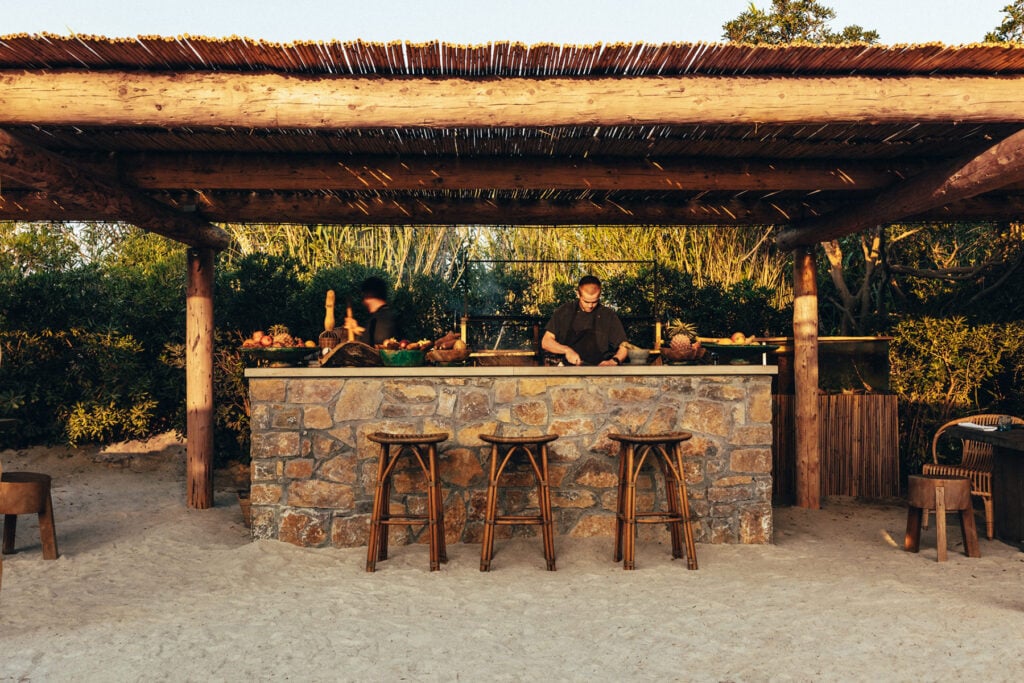 Epi 1959, Europe — Beachfront bar with stone counter, wooden stools, and thatch pergola at sunset, sand seating area with ambient lighting.