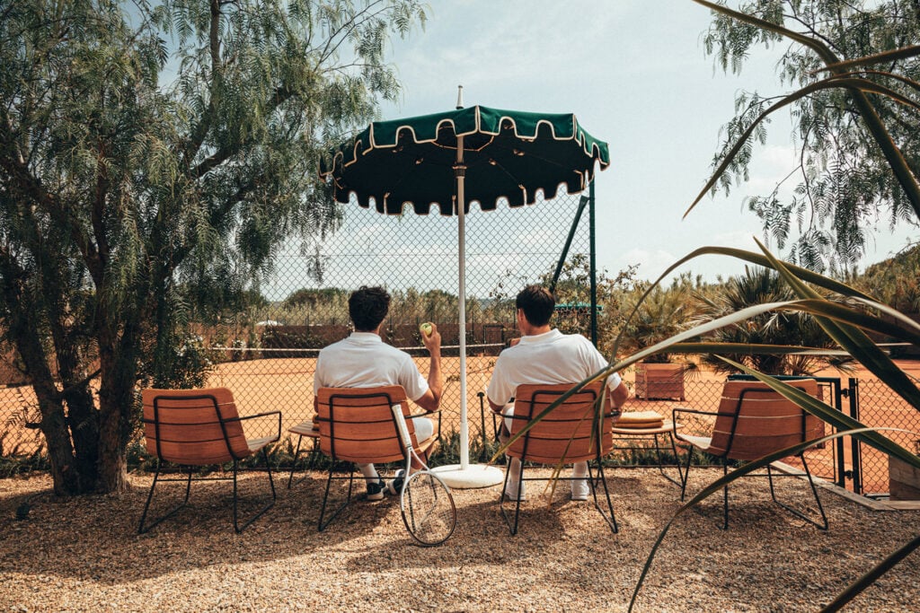 Epi 1959, Europe — Two figures in white shirts seated under green scalloped umbrella with terracotta chairs, desert landscape beyond mesh fence, weeping willow branches framing modernist poolside scene.