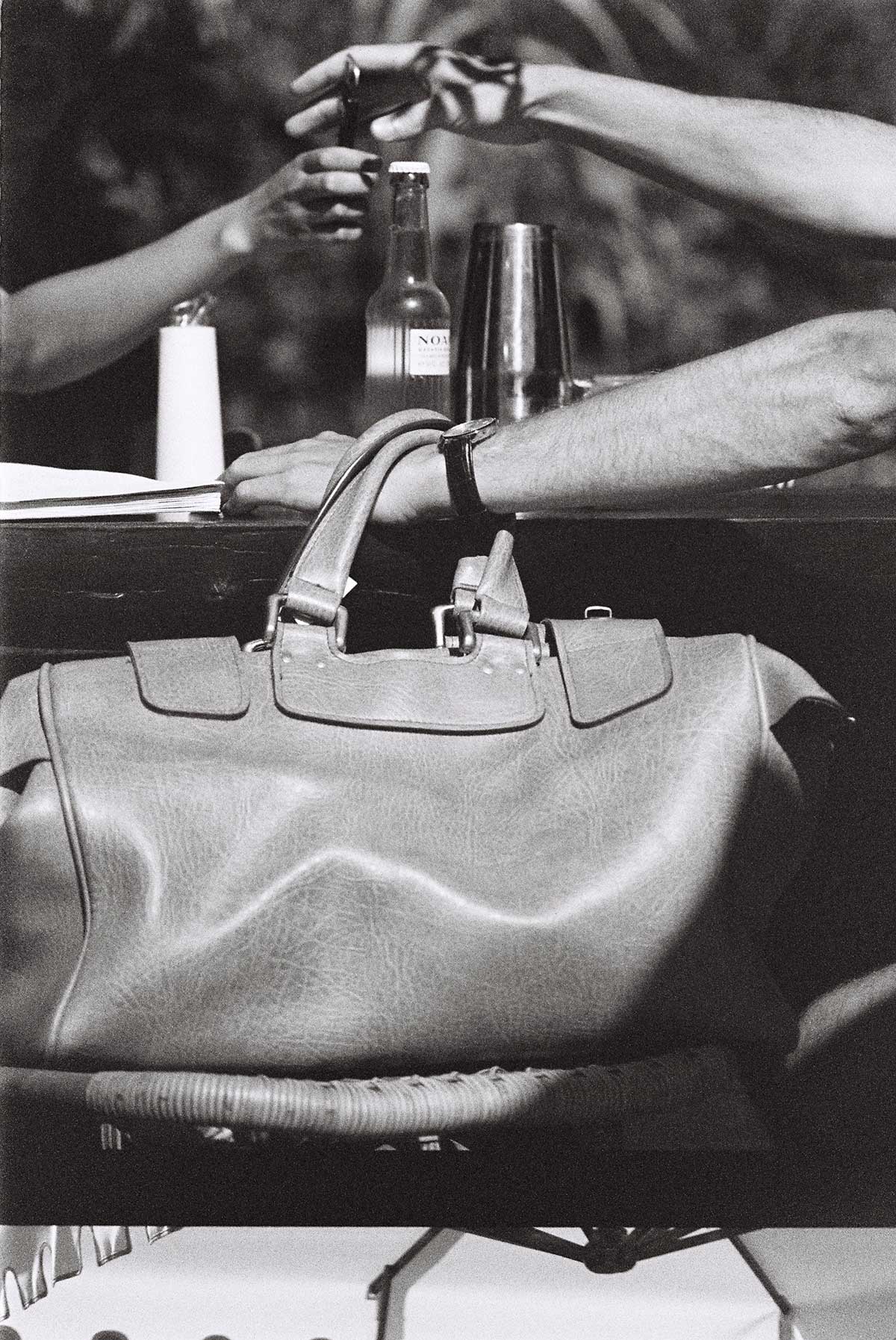 Epi 1959, Europe — Hands gesture over leather handbag and wine bottles in black and white editorial photography, luxury lifestyle aesthetic.