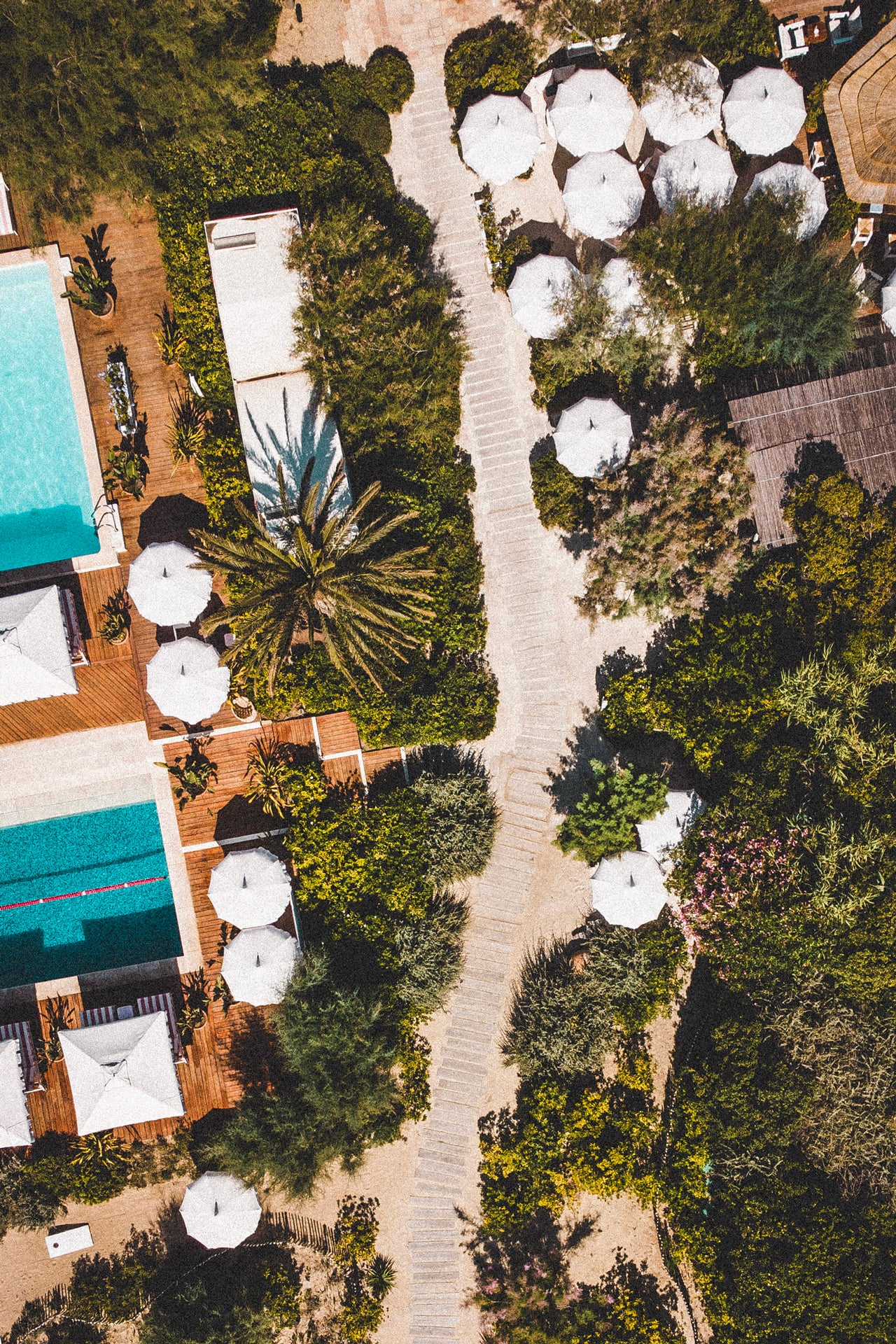 Epi 1959, Europe — Aerial view of poolside terrace with white umbrellas, wooden decking, lush landscaping, and stone pathways at luxury hospitality venue.